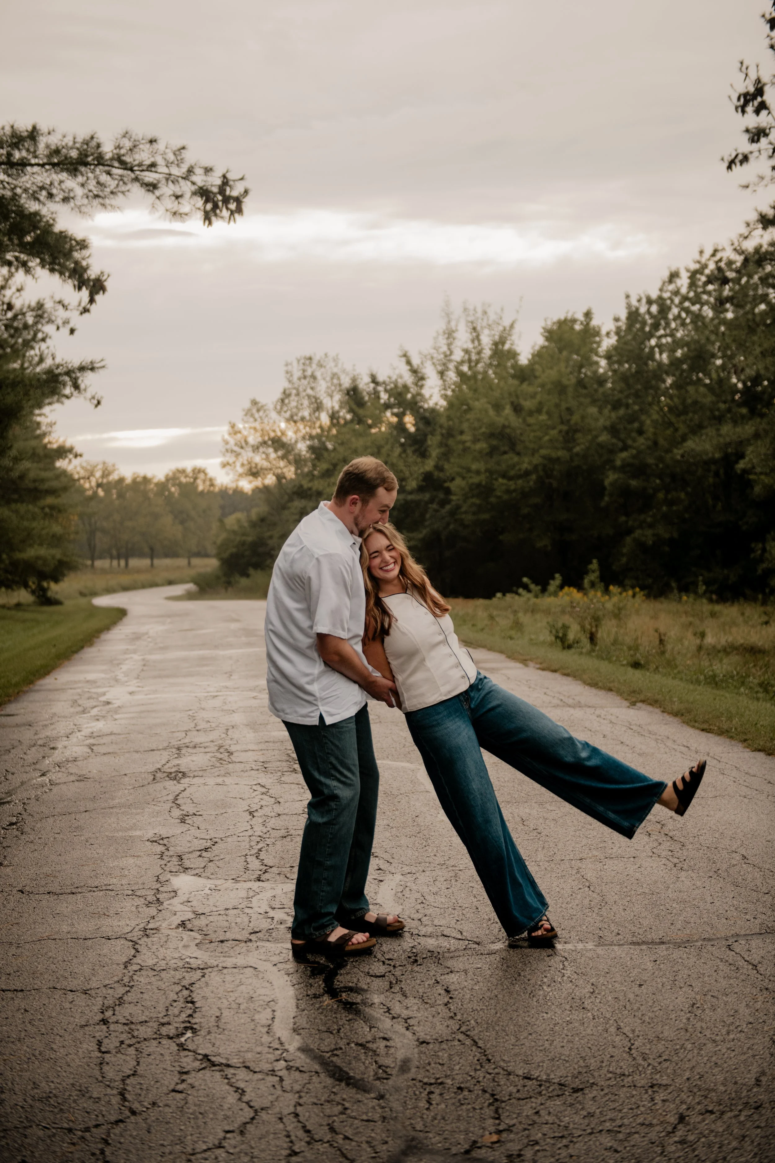  A picture of a young couple in the mid 20’s standing in the middle of a paved road at the Burr Oaks Conservation Area posing for an engagement portrait in the Kansas City Metro. The couple is being silly and dancing in the middle of the road. 