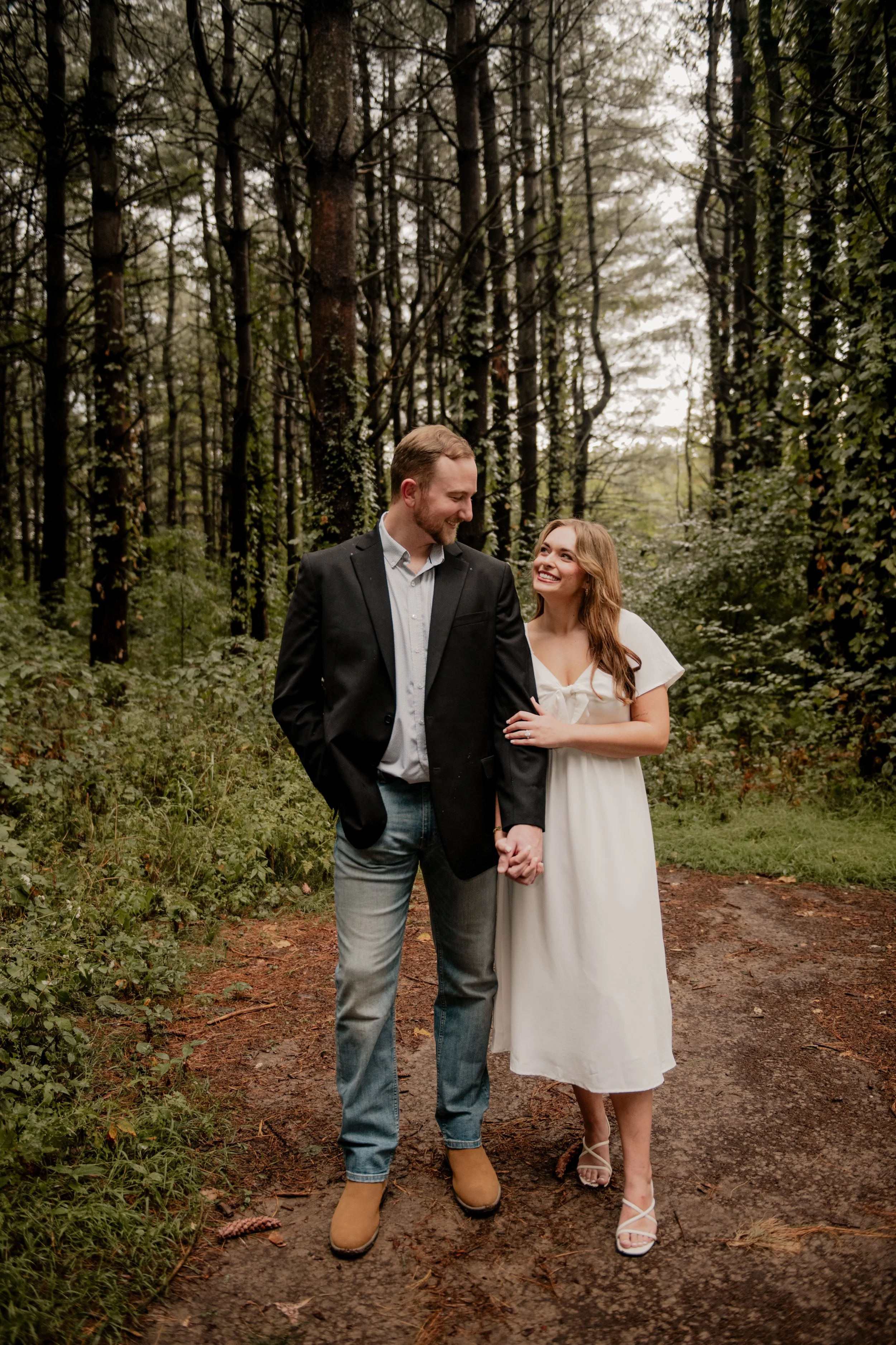  A pictured of a young couple in the mid 20’s standing in the middle of a forest at the Burr Oaks Conservation Area posing for an engagement portrait in the Kansas City Metro. The woman is holding the man’s hand and arm, snuggled up to each other smi