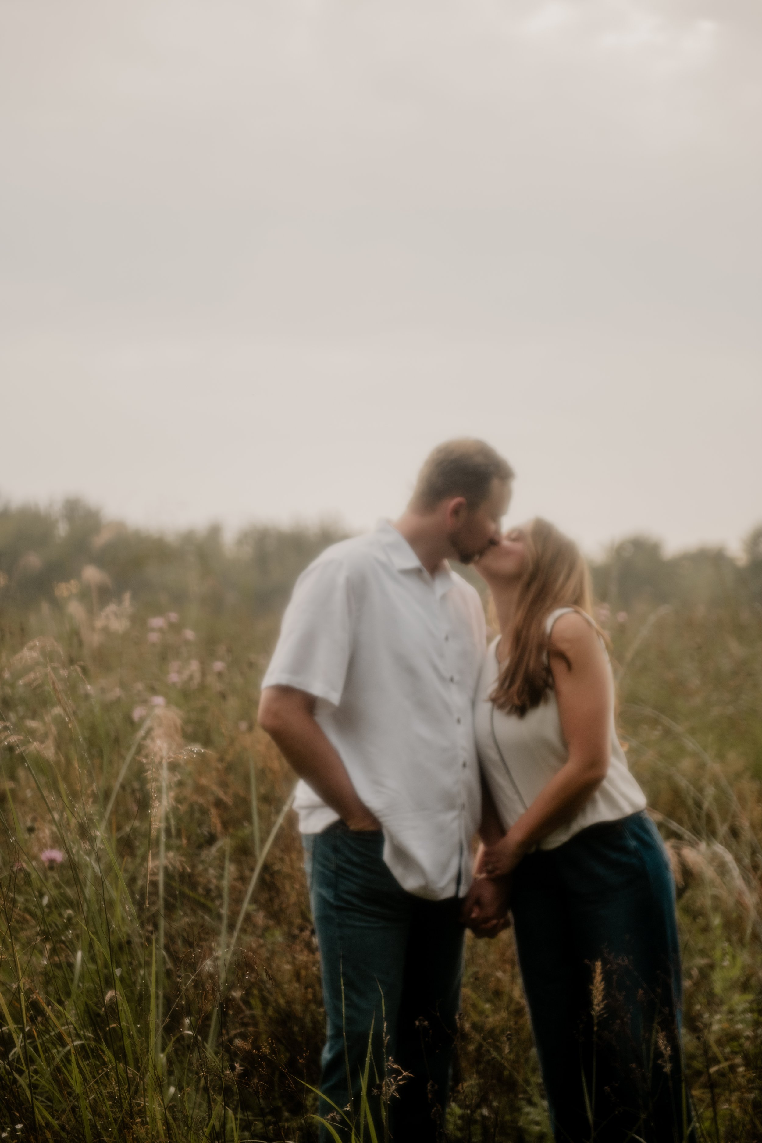  A pictured of a young couple in the mid 20’s standing an open field at the Burr Oaks Conservation Area posing for an engagement portrait in the Kansas City Metro. The couple share an intimate kiss. 