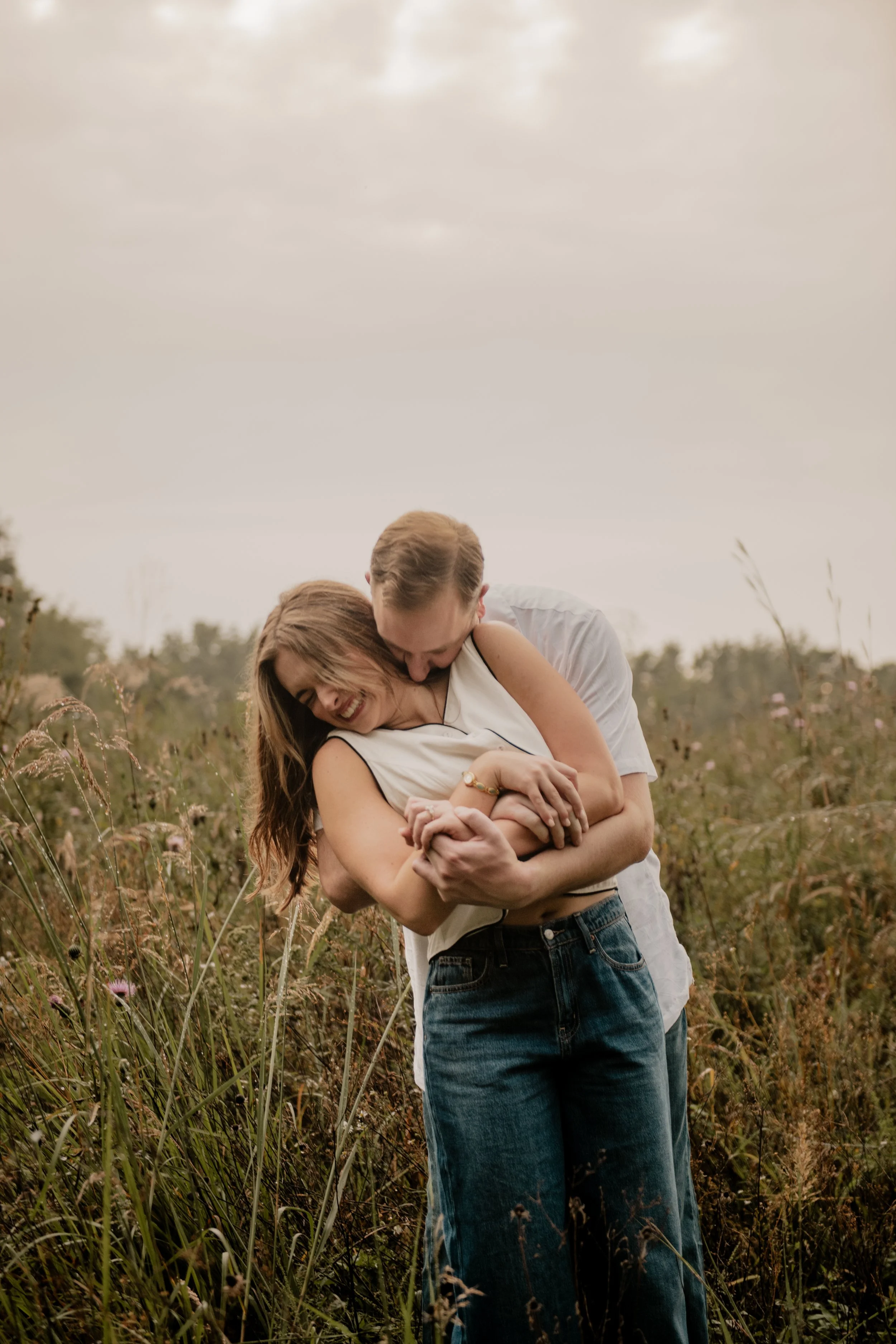  A pictured of a young couple in the mid 20’s standing in the middle of an open field at the Burr Oaks Conservation Area posing for an engagement portrait in the Kansas City Metro. The man hugs the woman from behind and kisses her collar bone, making