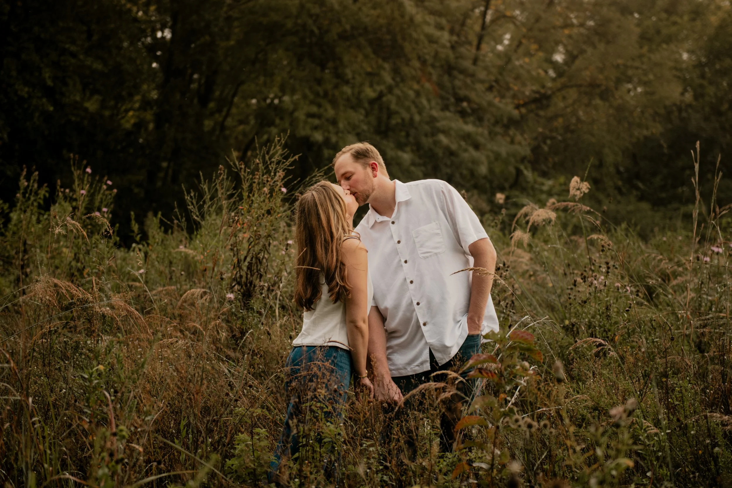  A pictured of a young couple in the mid 20’s standing in the middle of a field surrounded by trees at the Burr Oaks Conservation Area posing for an engagement portrait in the Kansas City Metro. The couple share an intimate kiss 