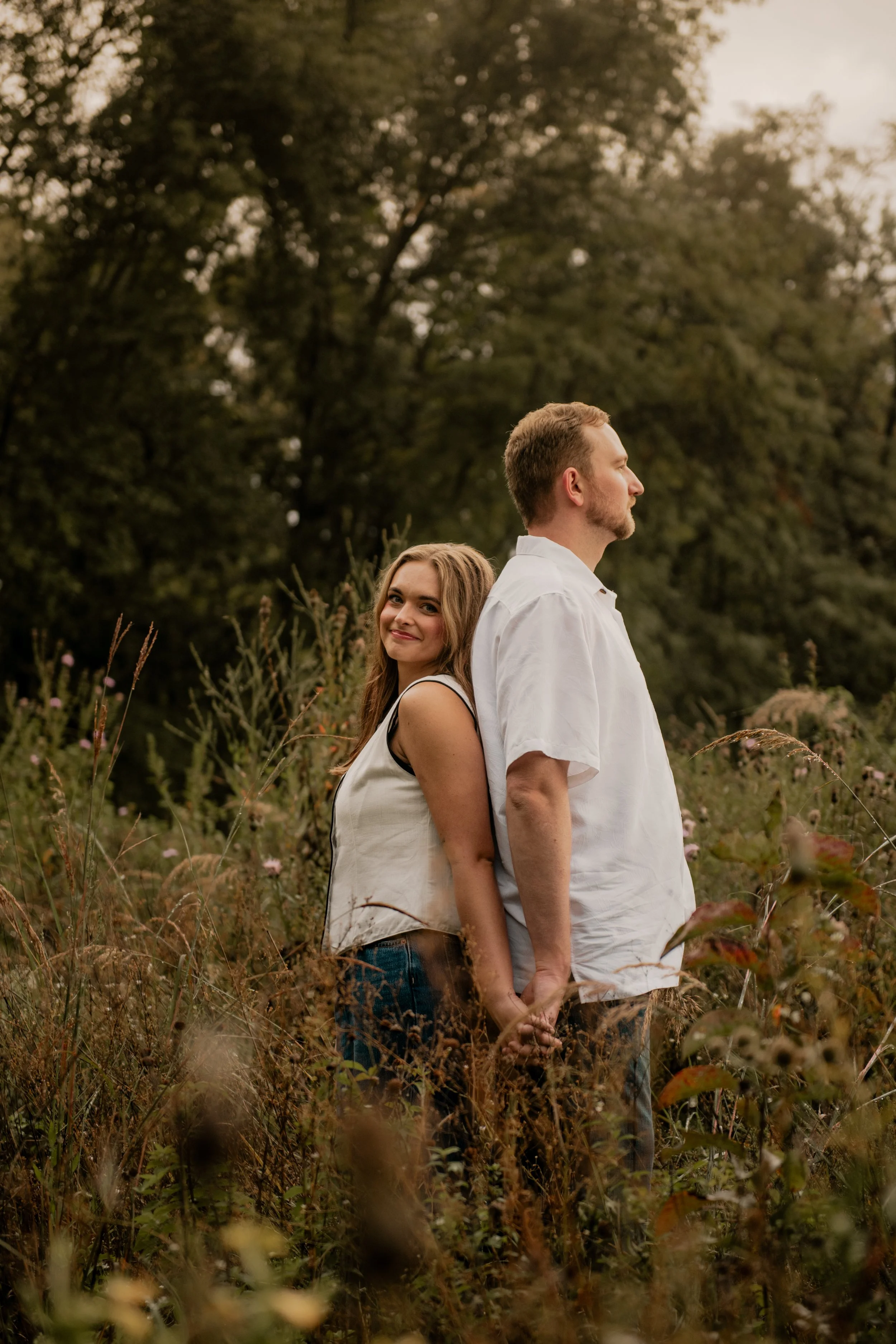 A pictured of a young couple in the mid 20’s standing in the middle of a field surrounded by trees at the Burr Oaks Conservation Area posing for an engagement portrait in the Kansas City Metro. The woman looks at the camera while the man stares off 