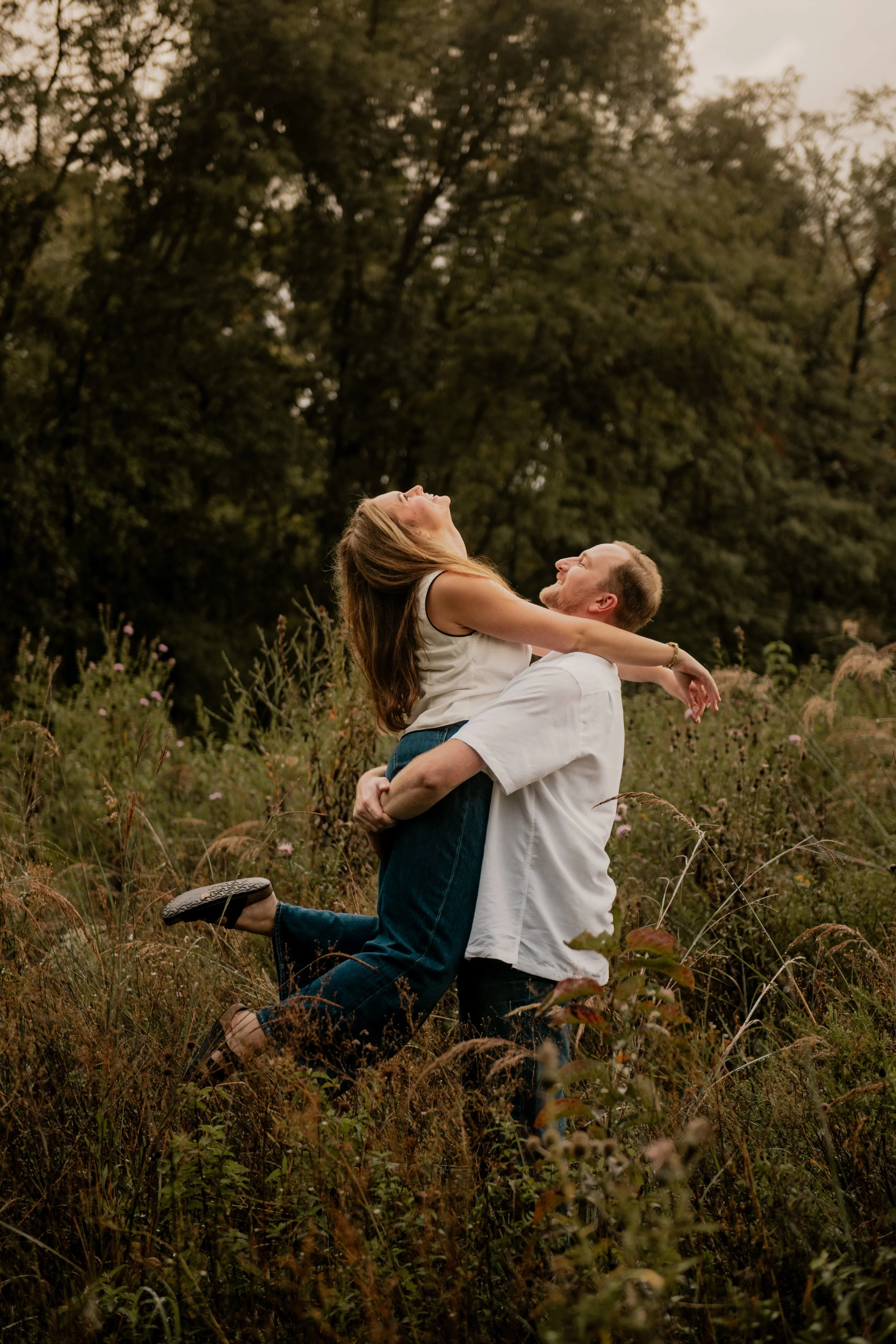  A pictured of a young couple in the mid 20’s standing in the middle of a field surrounded by trees at the Burr Oaks Conservation Area posing for an engagement portrait in the Kansas City Metro. The man picks up the woman and spins her around while s