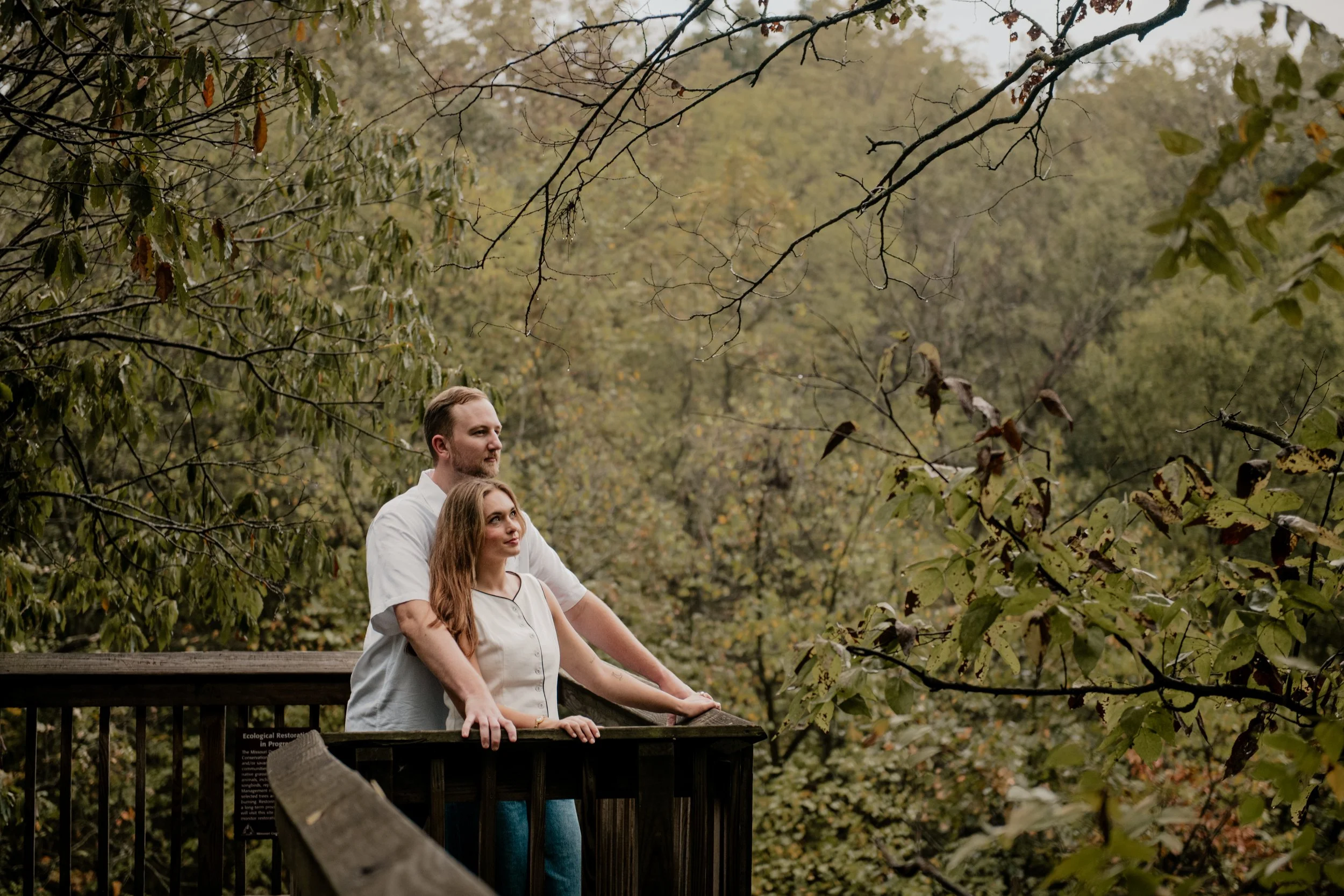  A picture of a young couple in the mid 20’s are standing on a wooden bridge at the Burr Oaks Conservation Area located in the Kansas City metro for engagement portraits. The couple stare off in the distance. 