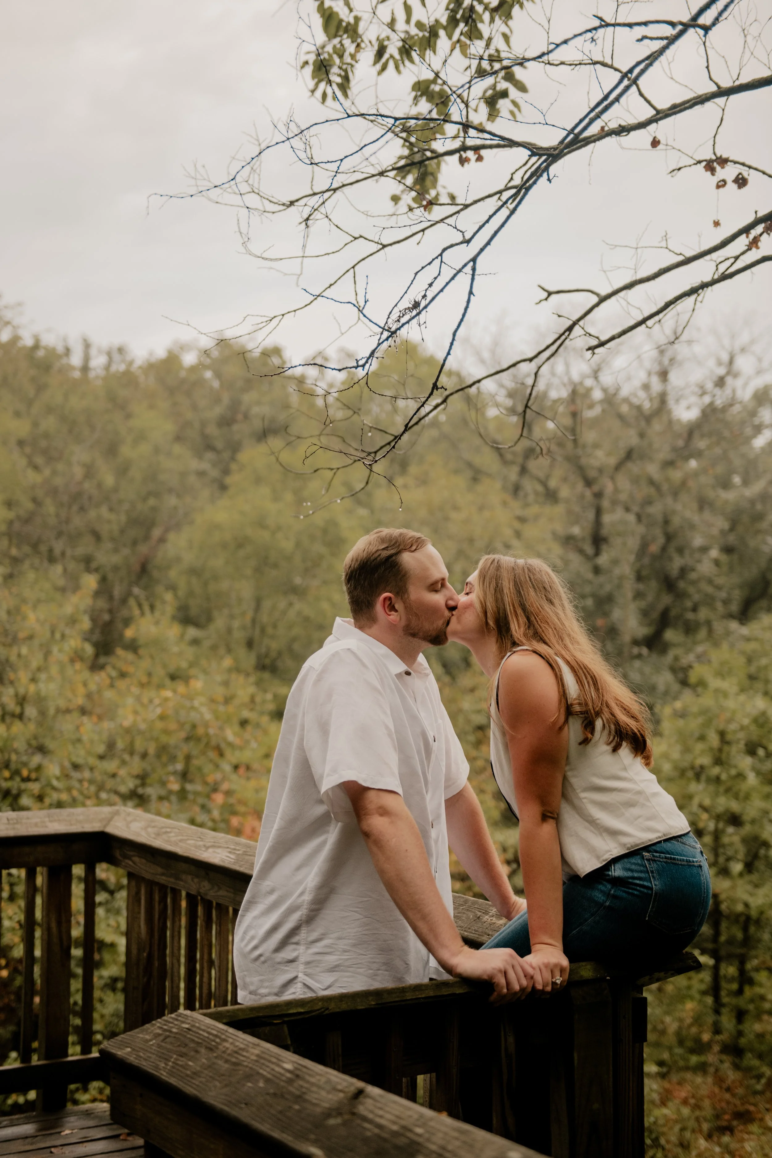  A picture of a young couple in the mid 20’s are standing on a wooden bridge at the Burr Oaks Conservation Area located in the Kansas City metro for engagement portraits. The woman sits on the railing of the bridge, and kiss the man. 