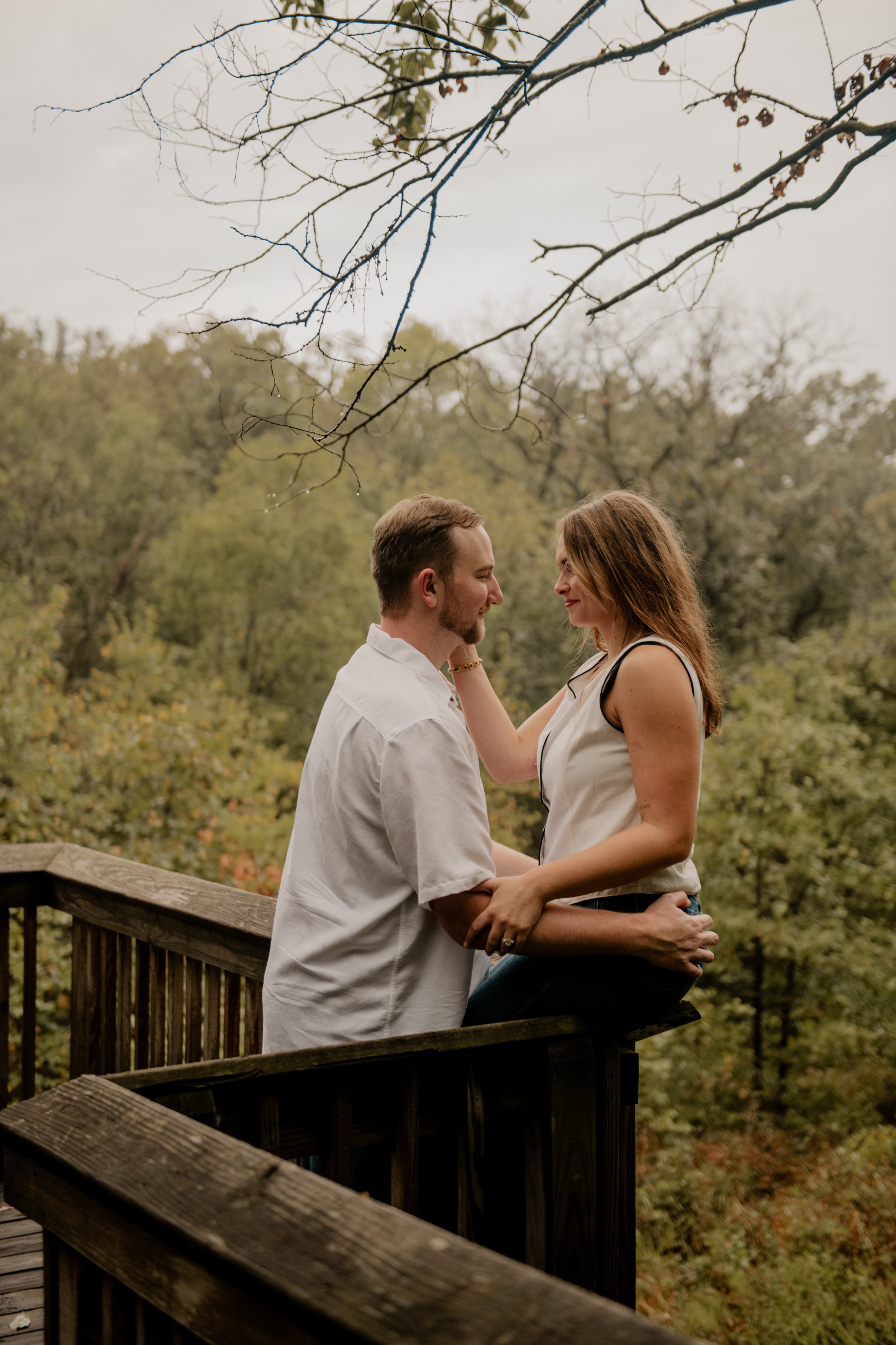  A pictured of a young couple in the mid 20’s are standing on a wooden bridge at the Burr Oaks Conservation Area located in the Kansas City metro for engagement portraits. The woman sits on the railing of the bridge and caresses the man’s face. 