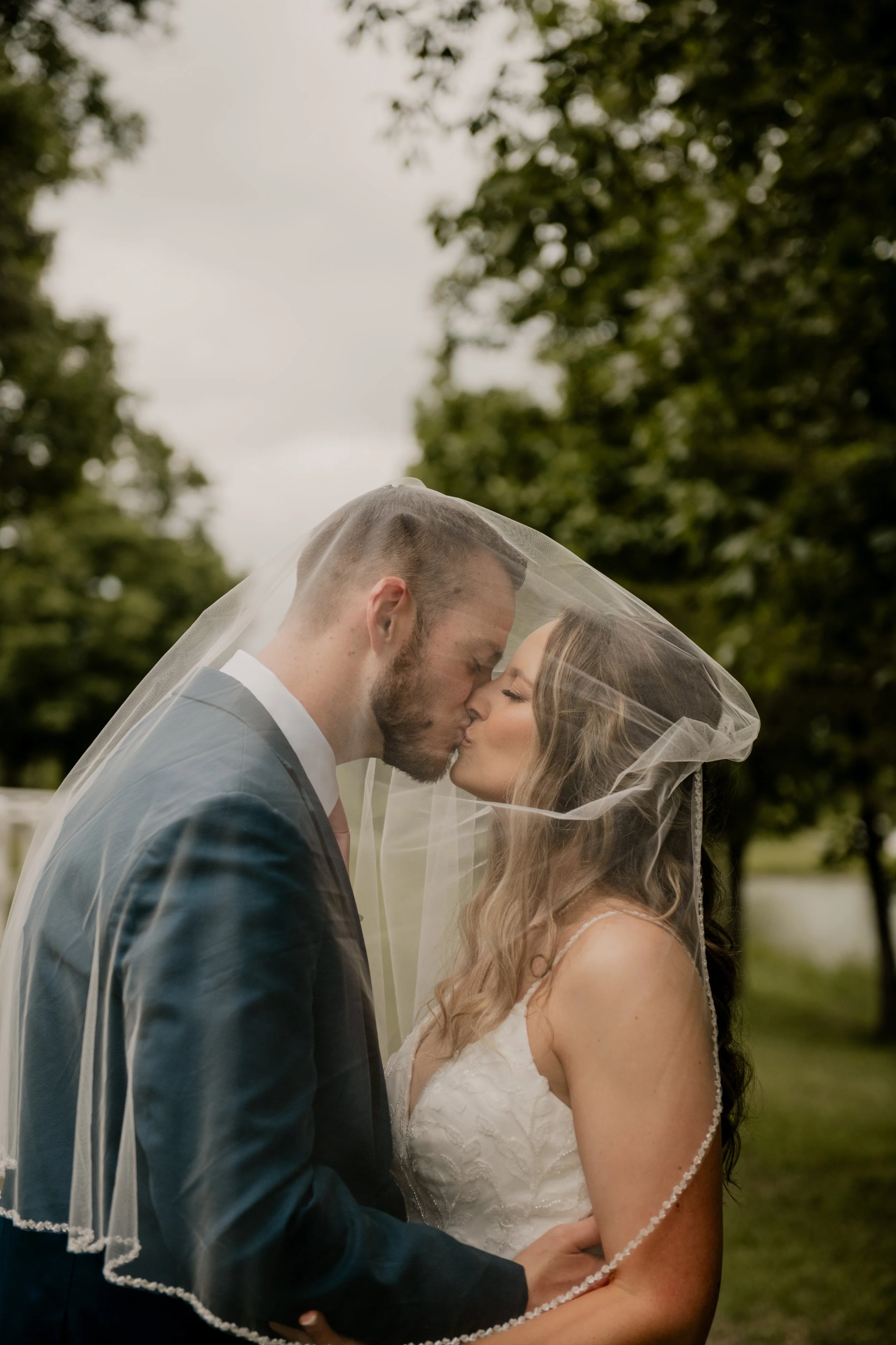 A bride and groom kiss under a veil outside with trees in the background.