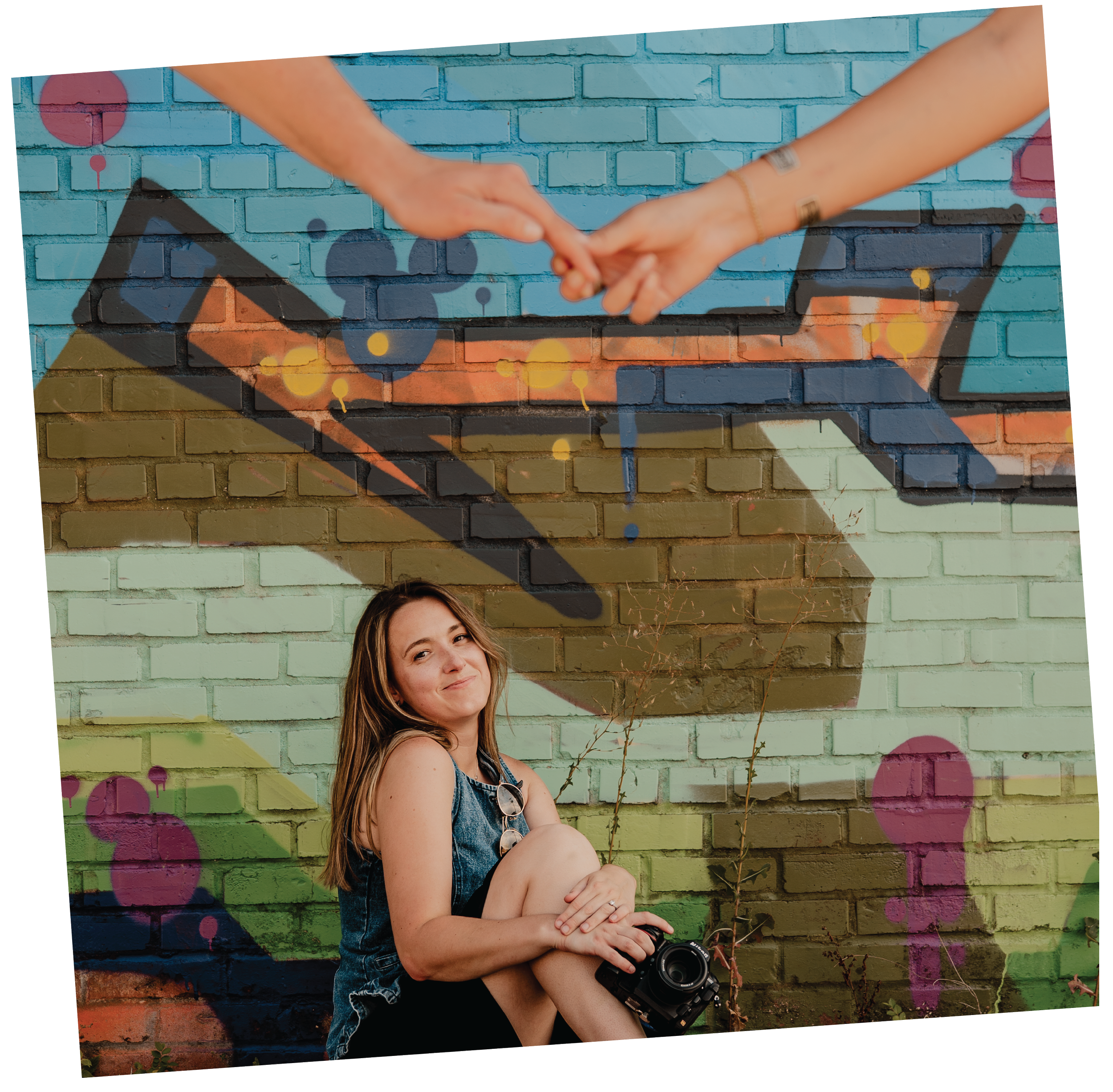 A woman sitting against a colorful brick mural, smiling and holding a camera. The mural features spray paint art with shapes, colors, and shadows, including a depiction of hands holding each other above her.