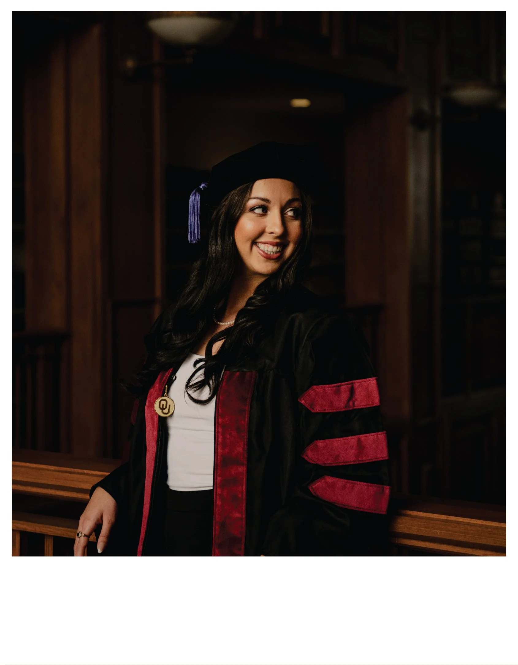 Young woman in scholarly regalia, wearing a cap and gown with red and black accents, smiling and looking to the side, standing in a wood-paneled library or academic setting.