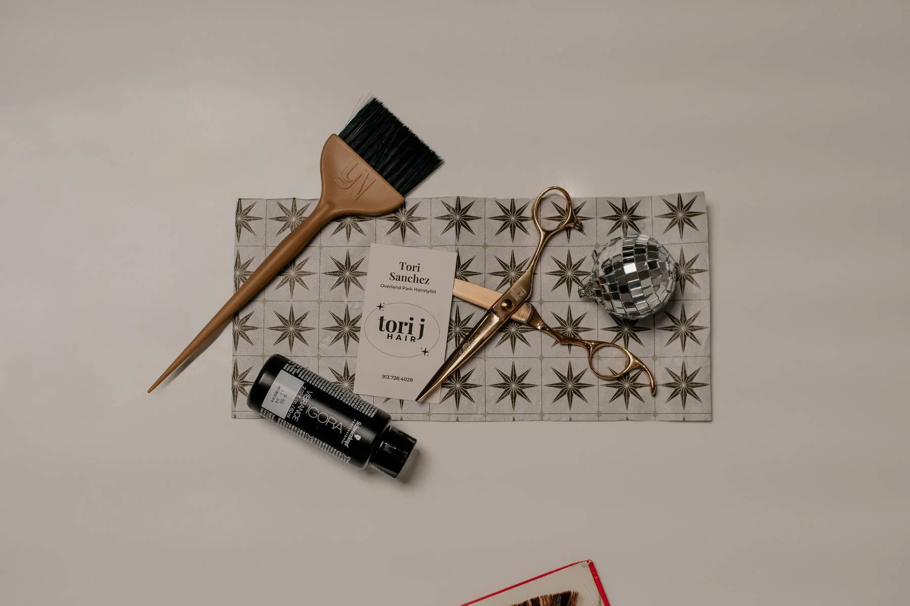 Hair styling tools on patterned paper, including a brush, scissors, a hair product, a disco ball, and a business card for Tori Sanchez, a hairstylist.