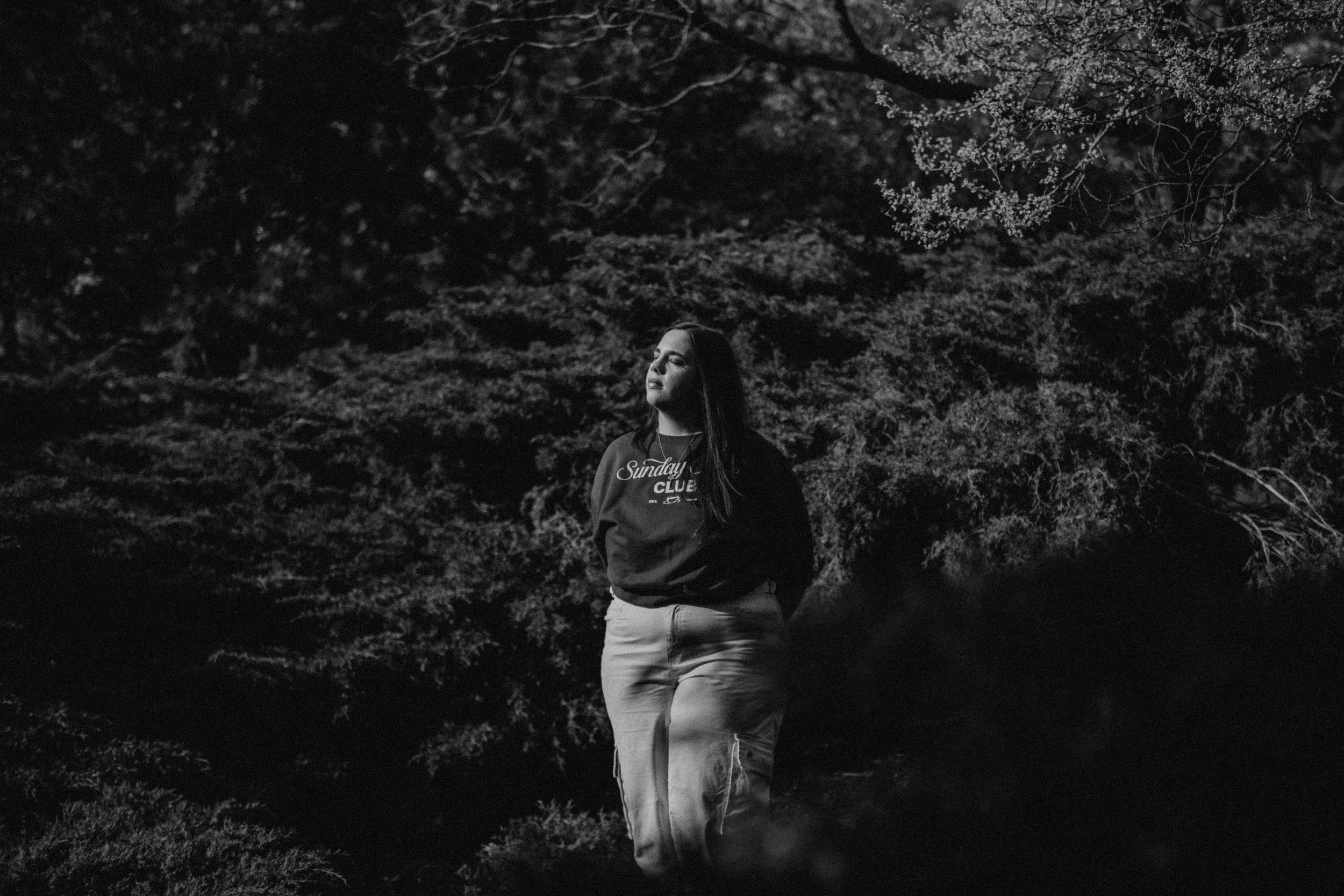 A woman standing outdoors at night in front of trees, with her eyes closed and her hands behind her back at the Nelson Atkins Museum of Art in Kansas City, Missouri.