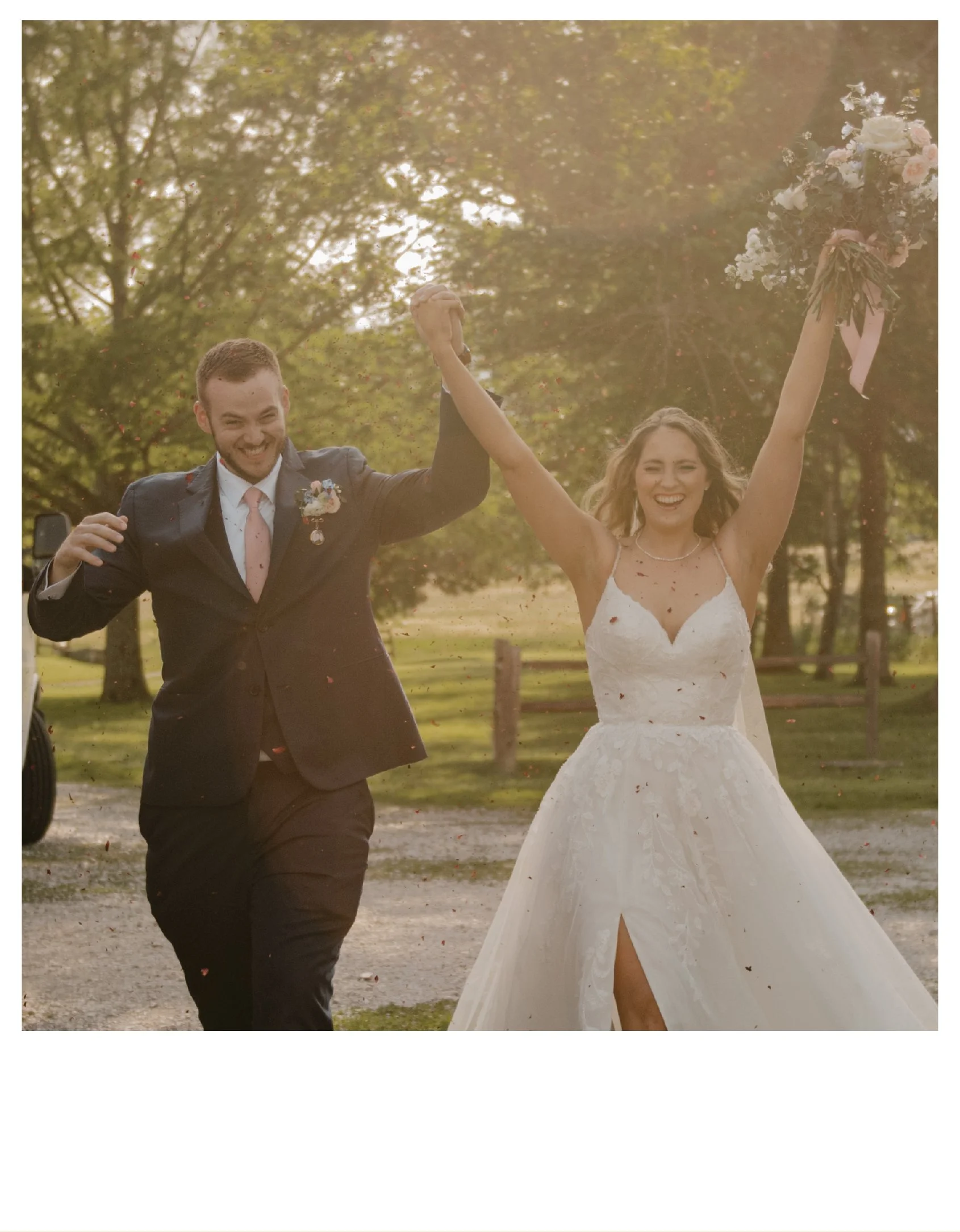 A newlywed couple celebrating outdoors, with the bride in a white wedding dress holding a bouquet and the groom in a dark suit, holding hands as they walk on a gravel path surrounded by trees at the Rogge Vineyard Raymore, Missouri.