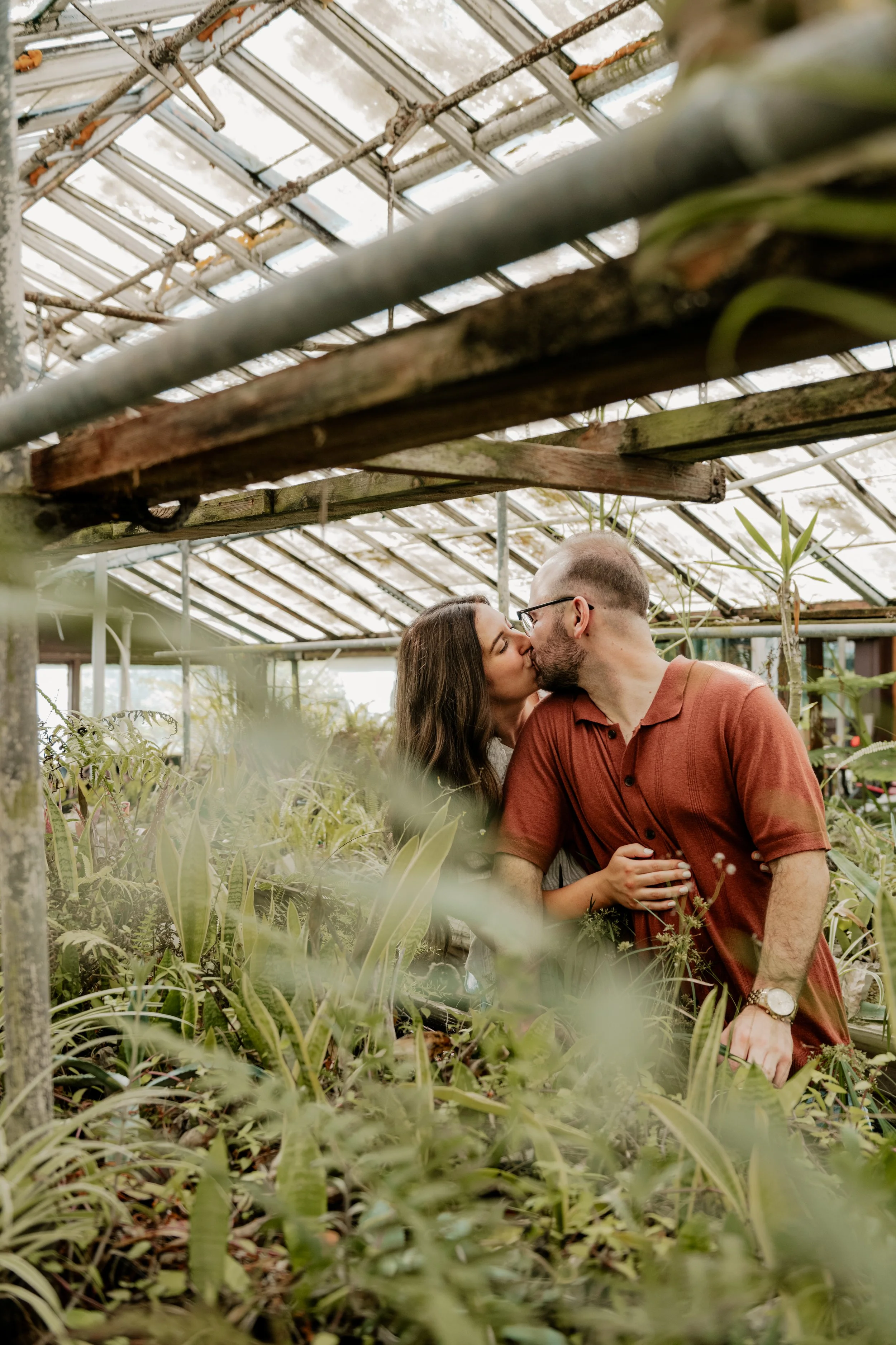 A couple sharing a kiss in a greenhouse filled with various green plants and foliage in John's Greenhouse in Kansas City, Missouri.