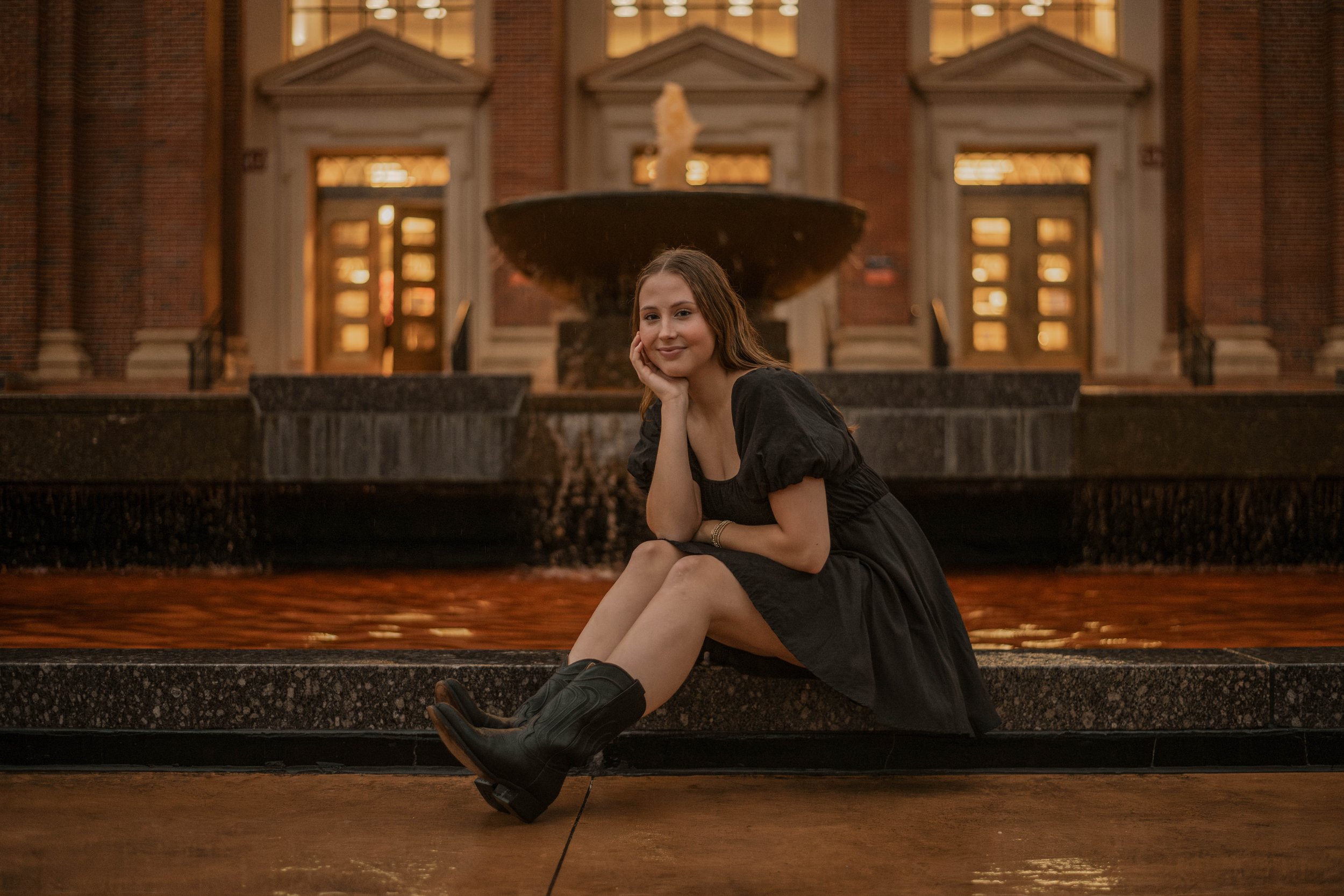 A young woman with long hair, wearing a black dress and cowboy boots, sitting on the edge of a fountain with her chin resting on her hand inside an ornate building with brick walls and illuminated windows at Oklahoma State University.