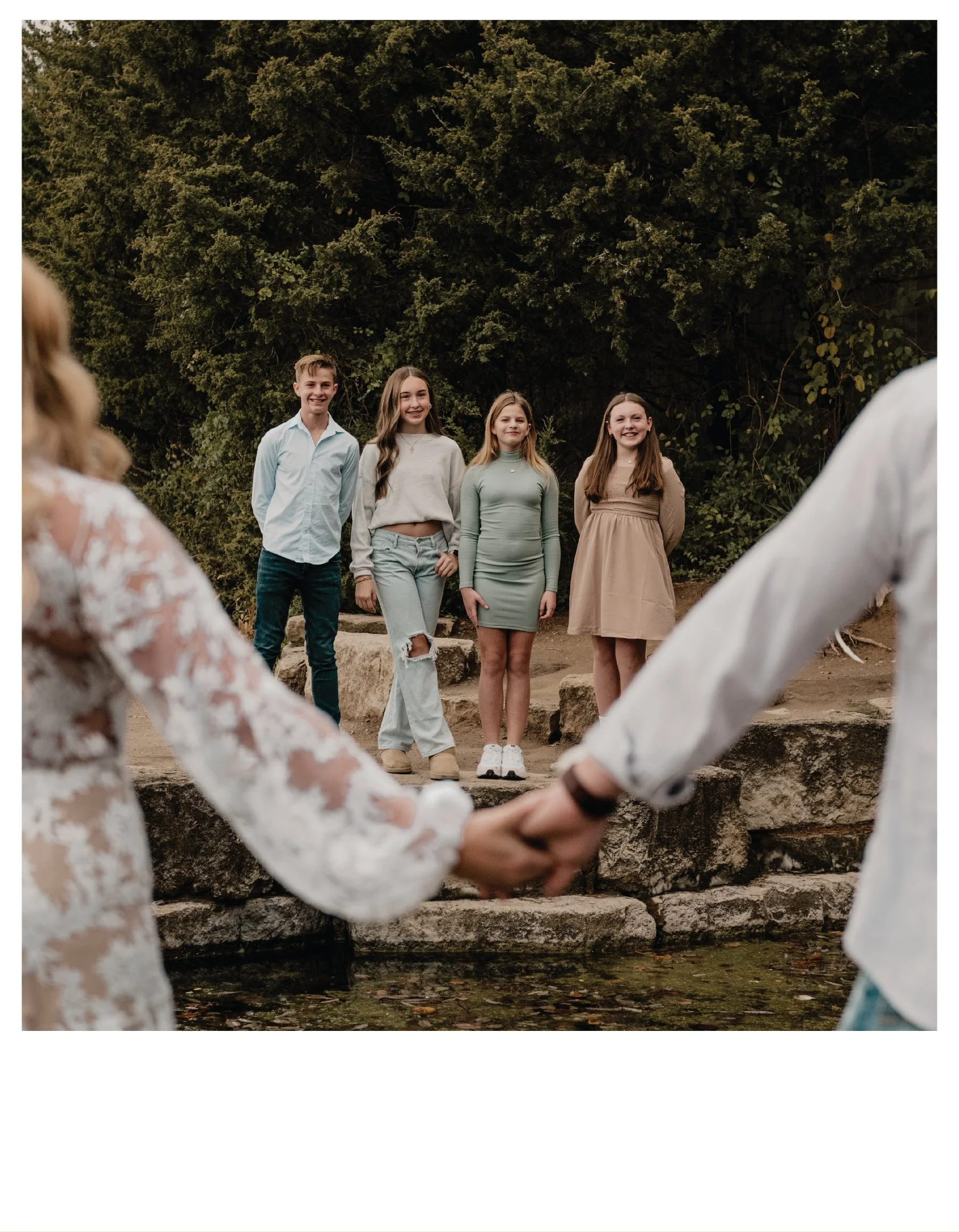 A couple holding hands near a creek with four people standing on rocks in the background, posing for a photo.