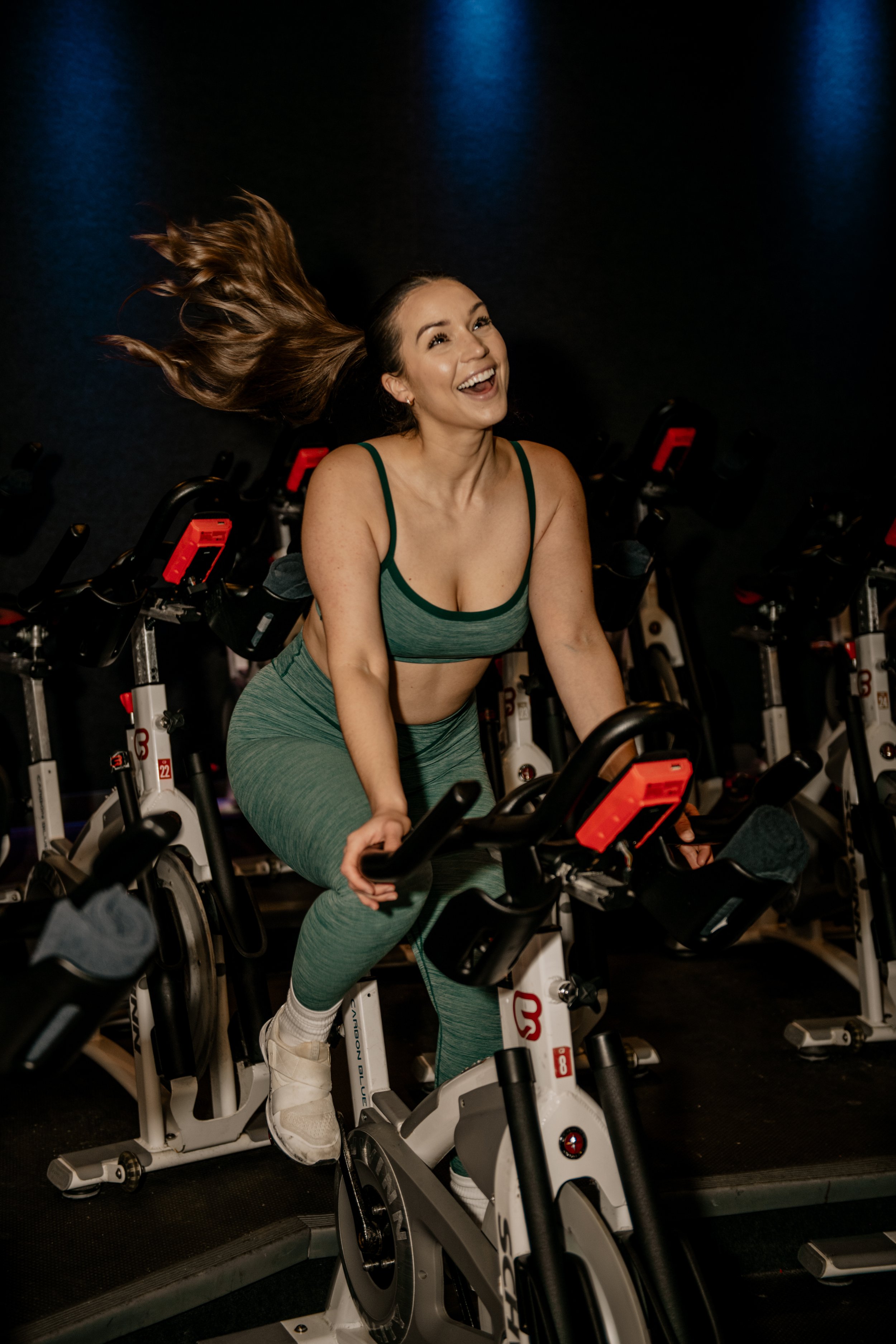 A woman with long hair riding a stationary exercise bike in a gym, smiling and laughing, surrounded by other exercise bikes.