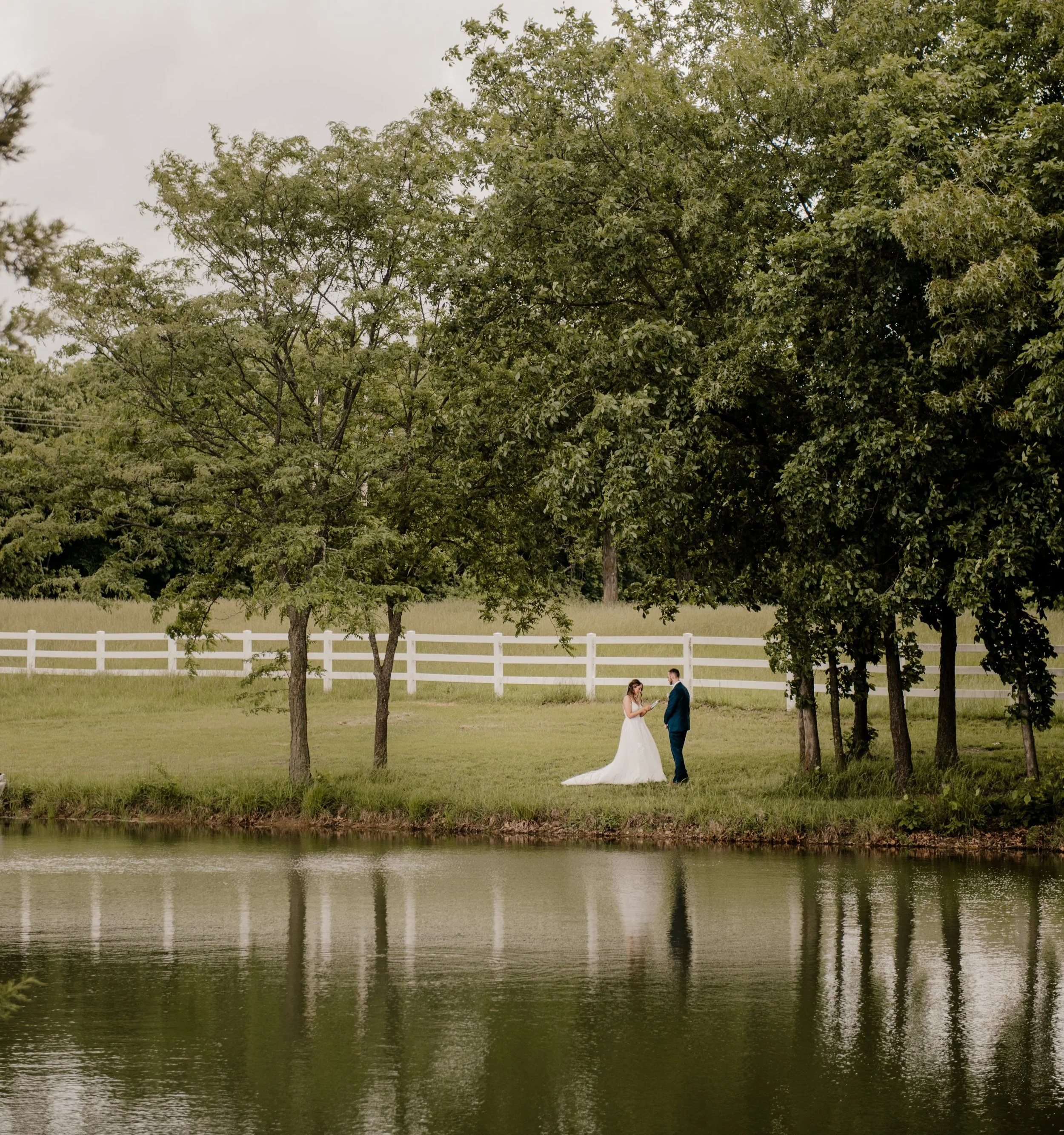 A bride and groom standing by a lake on their wedding day, surrounded by trees and a white fence in the background while they exchanged private vows.