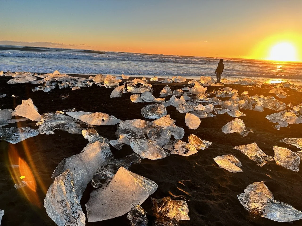 A person standing on a dark sandy beach at sunset, surrounded by large pieces of ice, with the ocean and the setting sun in the background.