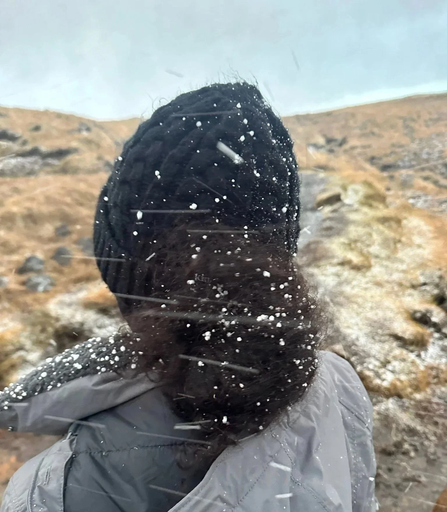 Person with long dark hair wearing a gray jacket standing outdoors in a windy, snowy hailstorm with Icelandic hills in the background.