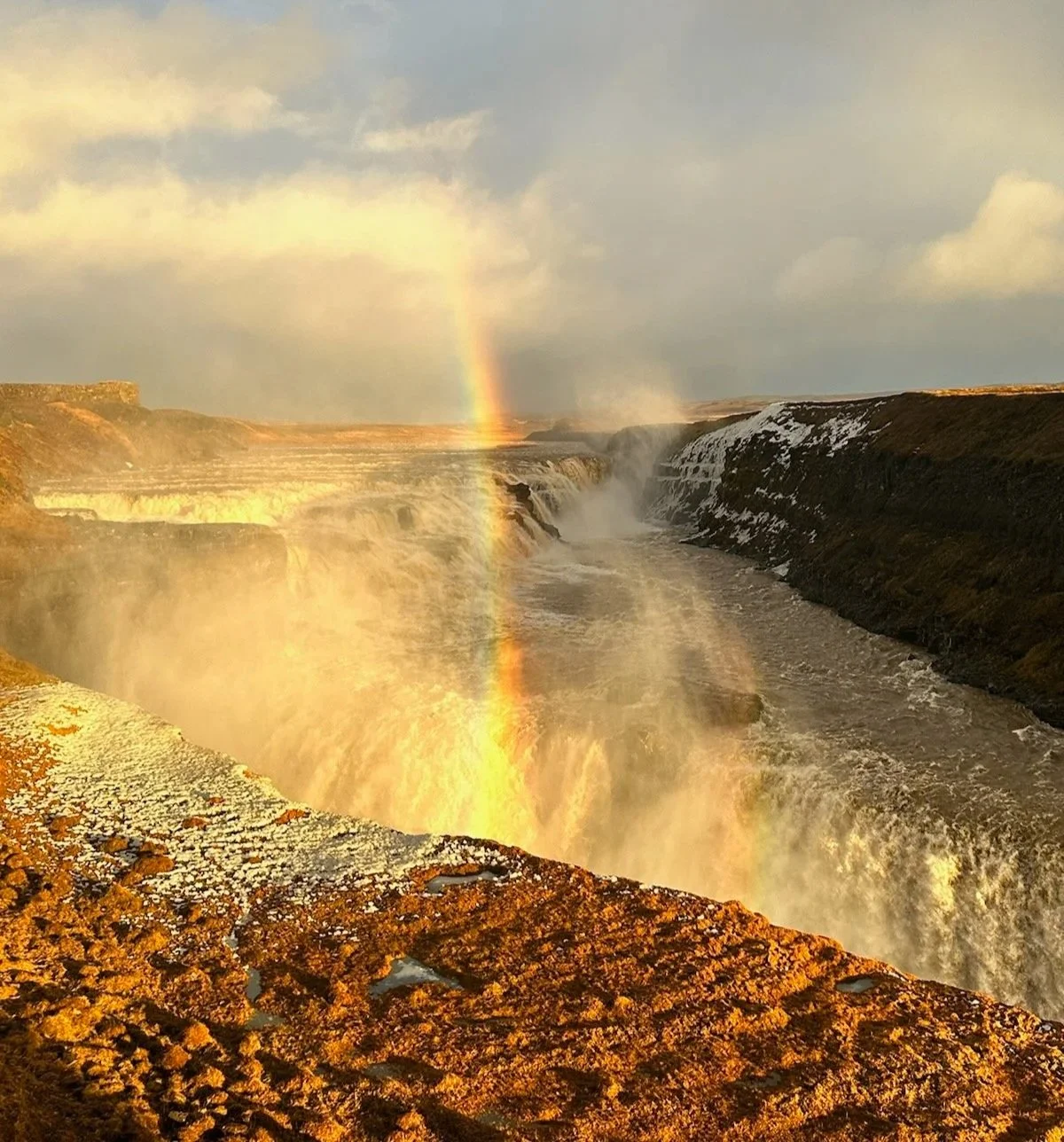 A waterfall with a rainbow and a small rainbow in the foreground, over a rocky ledge with moss and lichen, with clouded sky in the background.