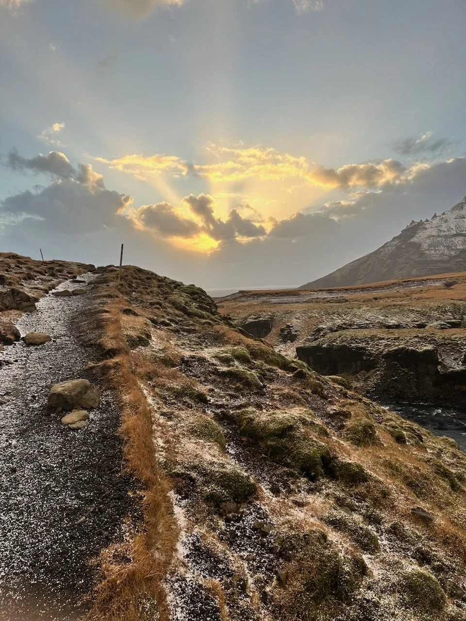 A rugged mountain trail with dirt and rocks, leading through a barren landscape with patches of snow or frost under a cloudy sky with sunlight breaking through.