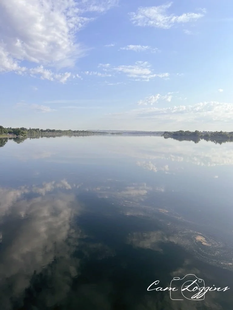 Calm river reflecting blue sky with scattered clouds, trees along the shoreline in the distance.