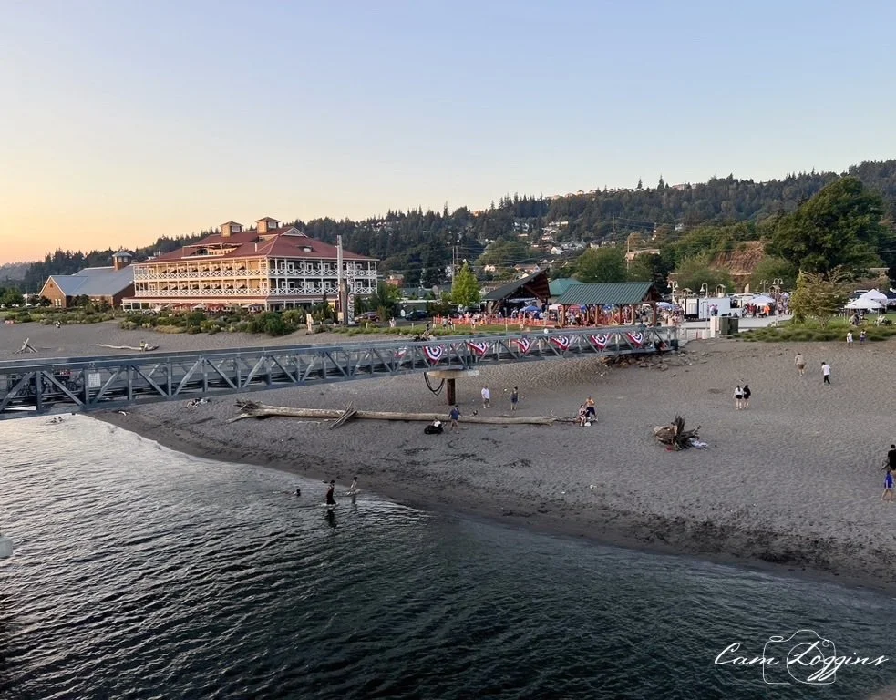 Beach with a metal walkway, people wading in the water, and a large building with red roofs in the background at sunset.
