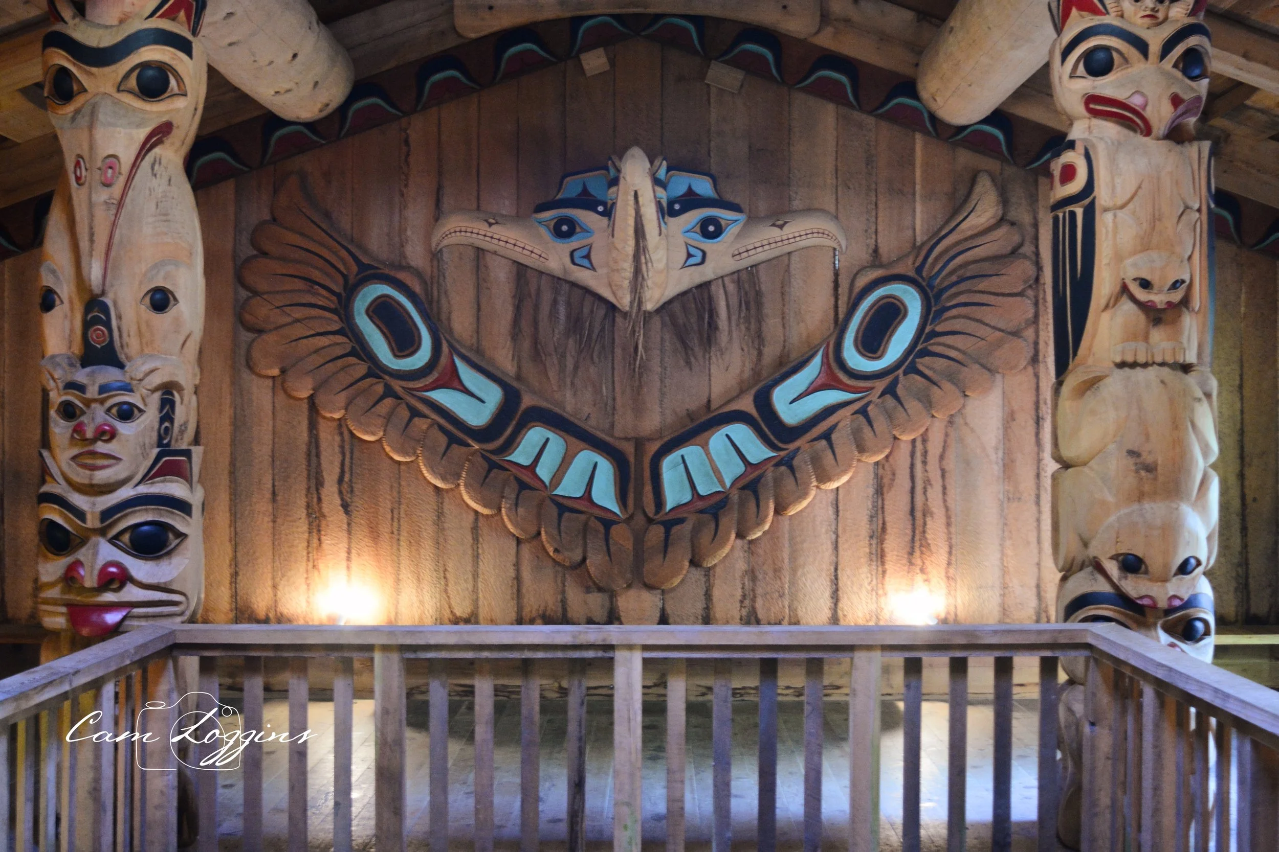 Interior of a Native American-style lodge with a large totem pole featuring various carved faces and animal shapes, including a butterfly with eyes, surrounding a wall with a carved face and winged figure.