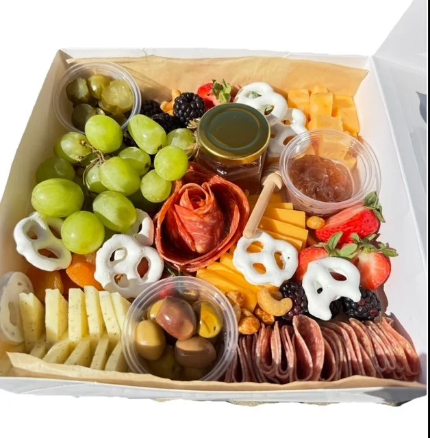 A cheese and fruit platter with grapes, strawberries, assorted cheeses, cured meats, crackers, honey jar, olives, and various garnishes arranged in a white box.