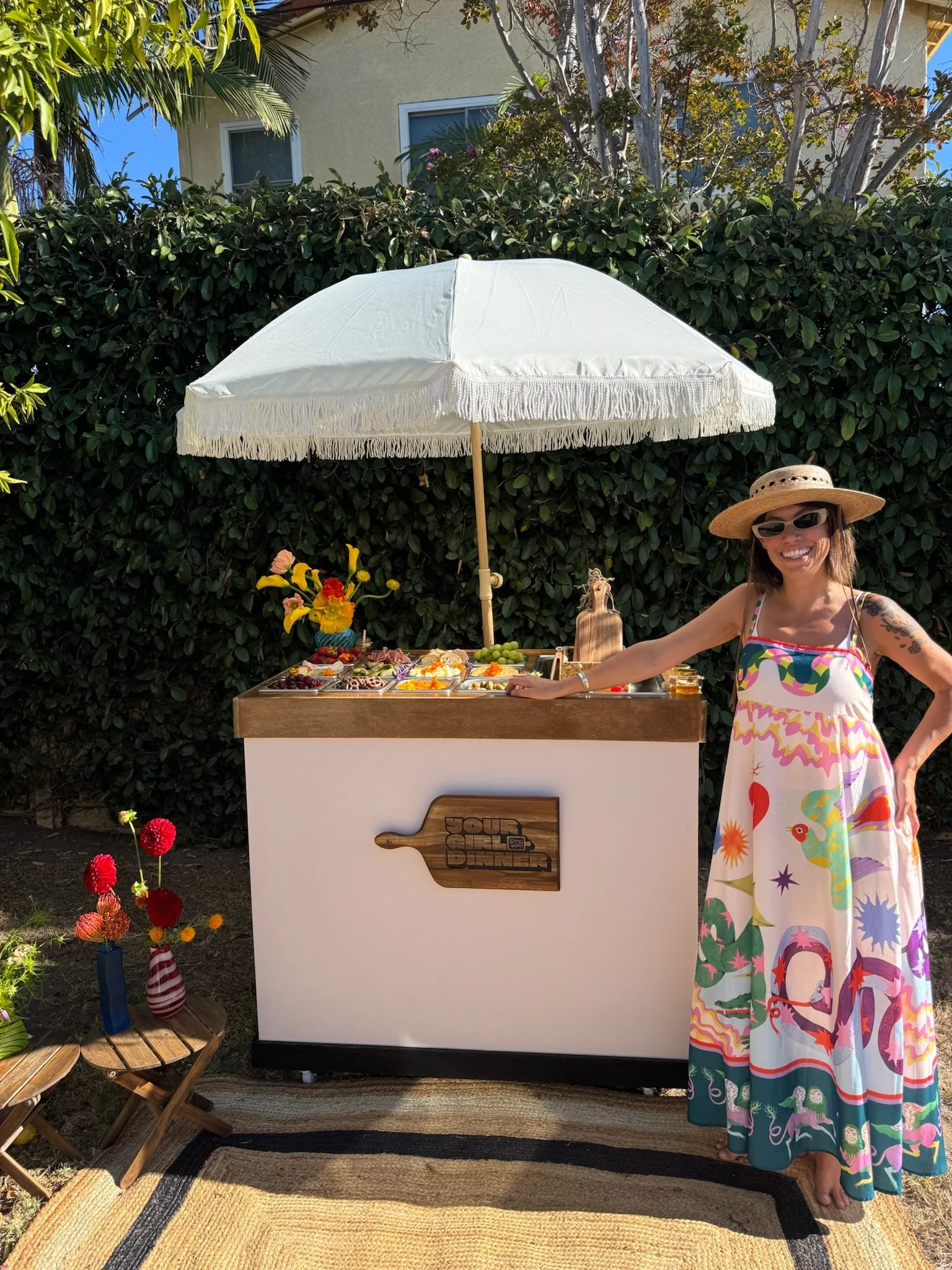 Woman smiling in a colorful dress and wide-brimmed hat standing next to a portable food stand with an umbrella, surrounded by greenery and small flower arrangements.