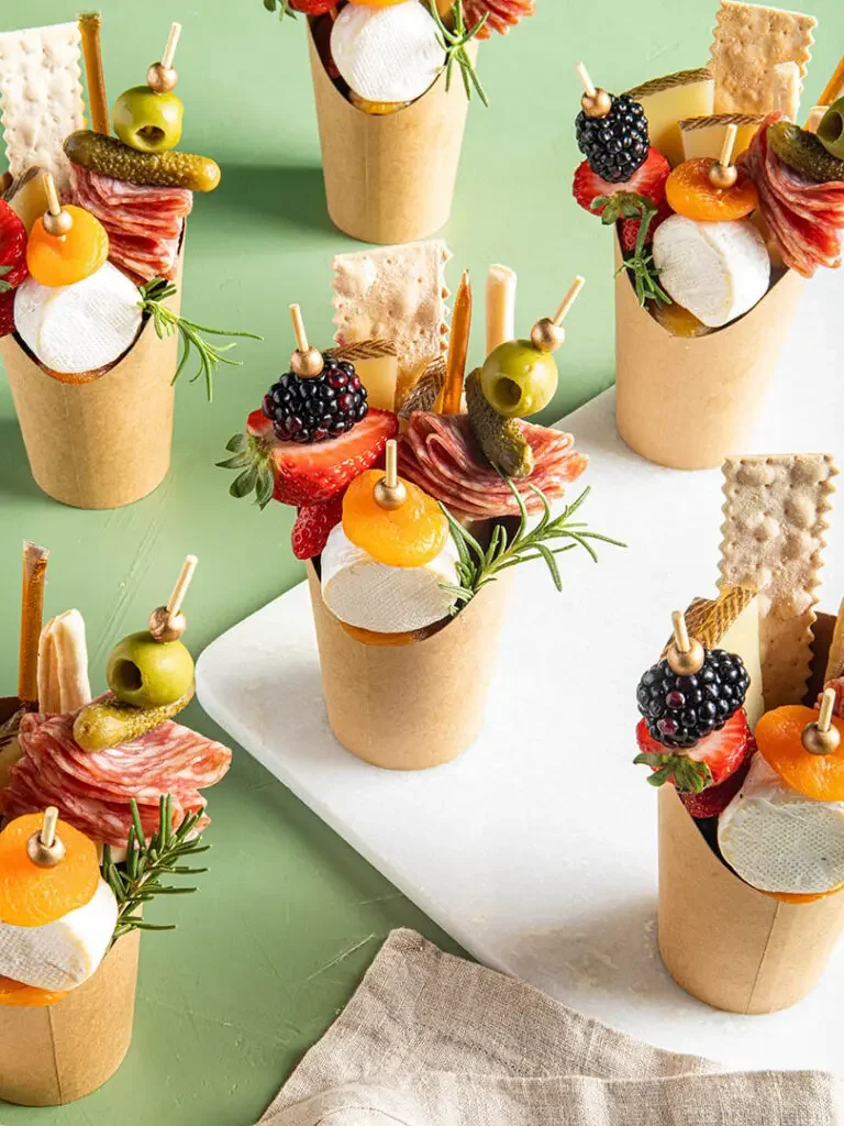 An assortment of individual charcuterie cups filled with cheese, cured meats, fresh fruits, crackers, and garnished with herbs, arranged on a white board and green background.