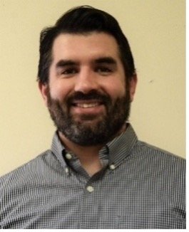 A man with dark hair and a beard, wearing a checkered button-up shirt, smiling at the camera against a plain light-colored background.