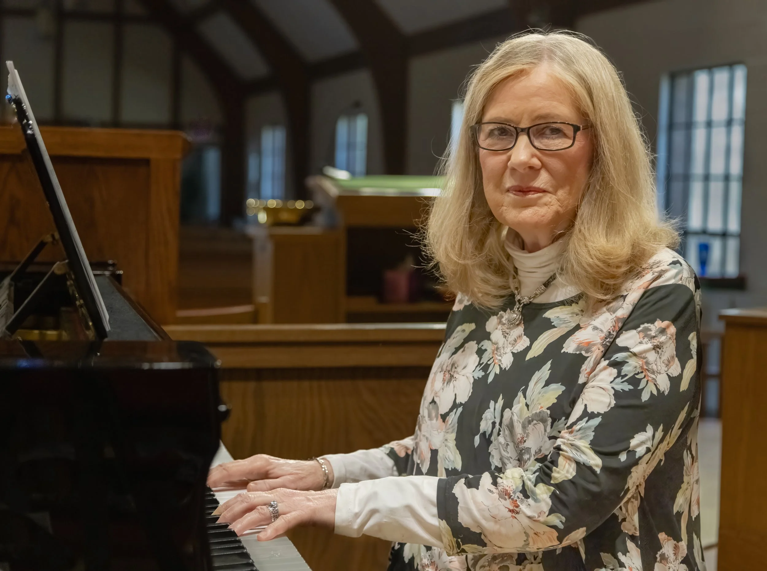 An older woman with glasses and blonde hair, wearing a floral-patterned jacket, standing next to a black Yamaha piano in a church or worship space.