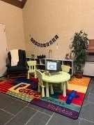 Children's playroom with small chairs around a table, a TV on a stand, a colorful rug, and a plant in the background.