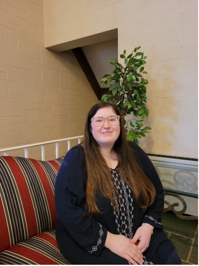 A woman with long brown hair, glasses, and a black sweater sitting on a striped red, black, and beige bench indoors, with a large green plant and a beige brick wall in the background.