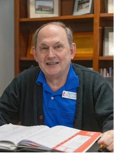 A smiling man with short hair wearing a blue polo shirt and a dark cardigan, seated at a desk with an open book, in front of a wooden bookshelf with framed pictures.