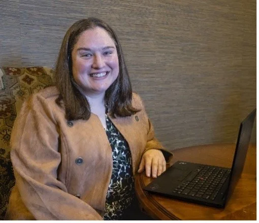 A smiling woman with shoulder-length brown hair sitting at a round wooden table with a laptop in front of her.