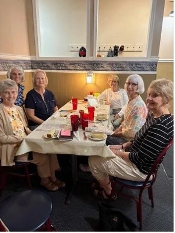 Seven women sitting around a dining table with food and drinks in a room with a mirror and window.