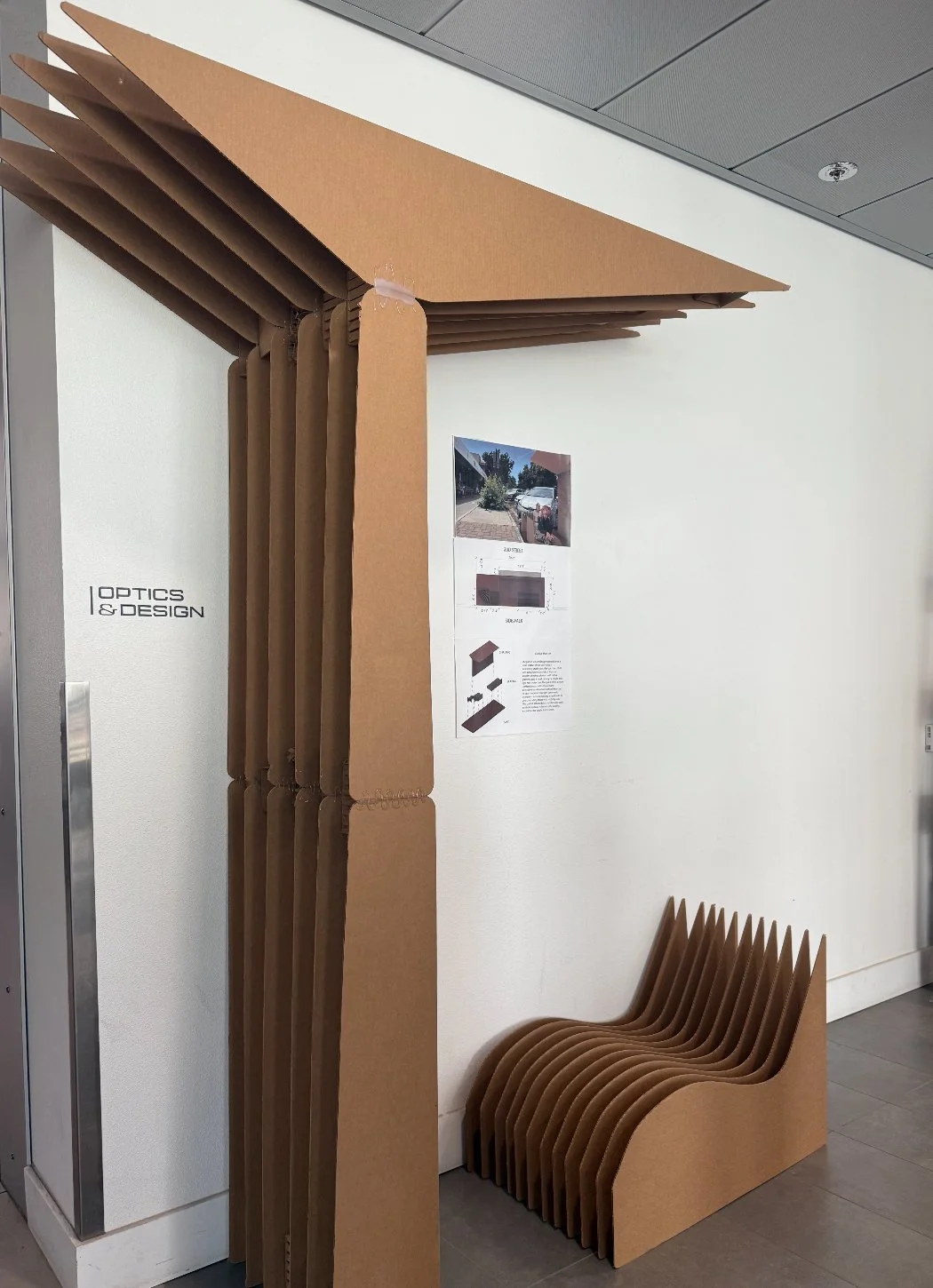 A stack of corrugated cardboard pieces forming the shape of a chair, placed against a white wall with a seating area made from similar cardboard material.