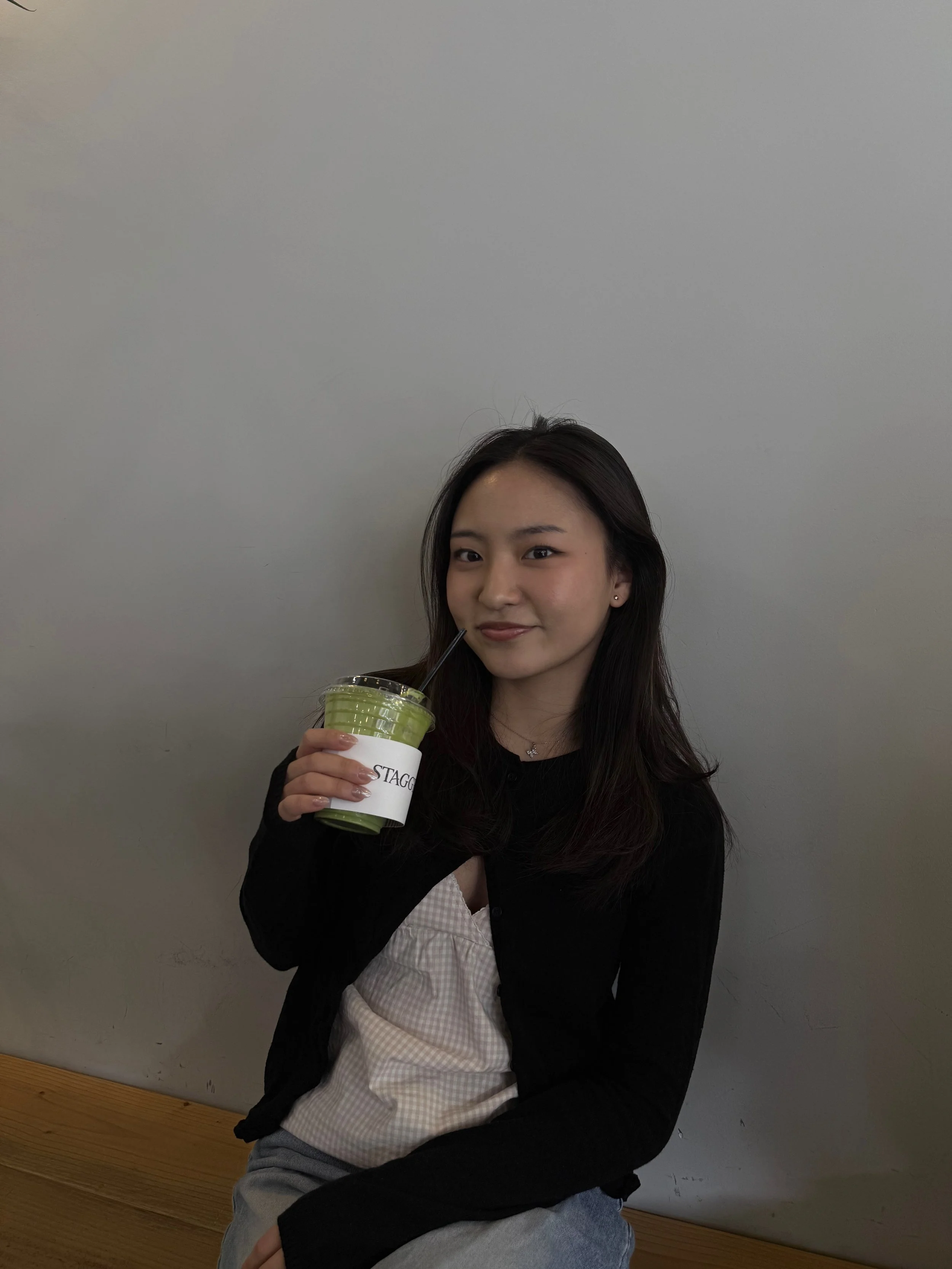 Young woman sitting on a wooden bench, holding a green smoothie with a black straw, smiling at the camera, against a plain light gray wall.