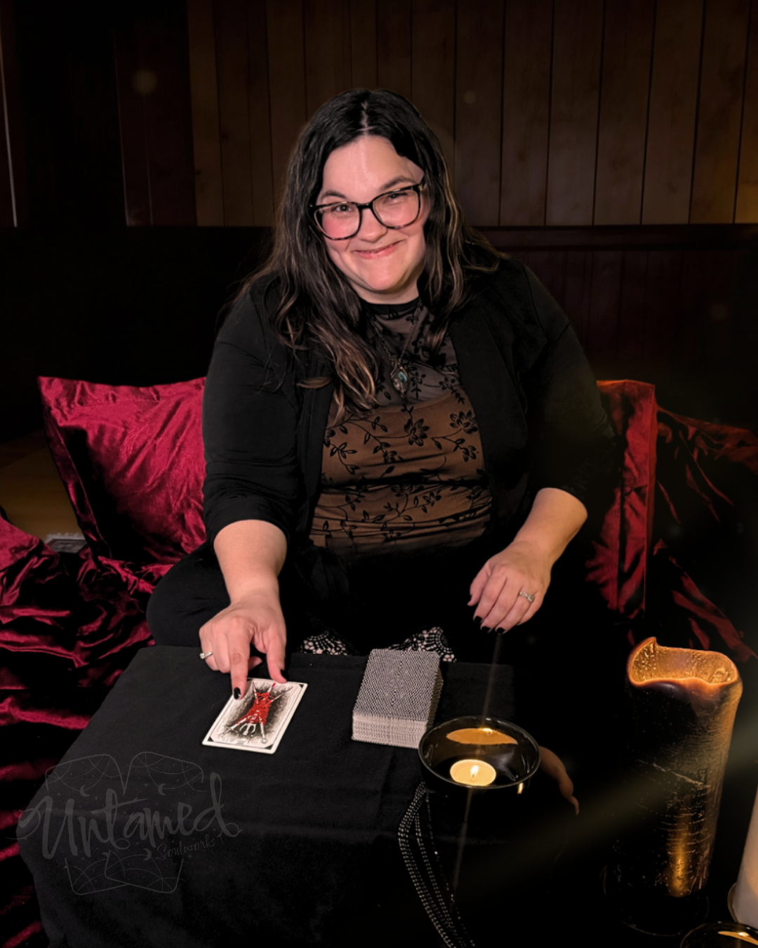 A woman with glasses and long dark hair smiling while sitting on a black couch with red pillows. She is holding a tarot card and has a deck of tarot cards and candles on a black table in front of her.