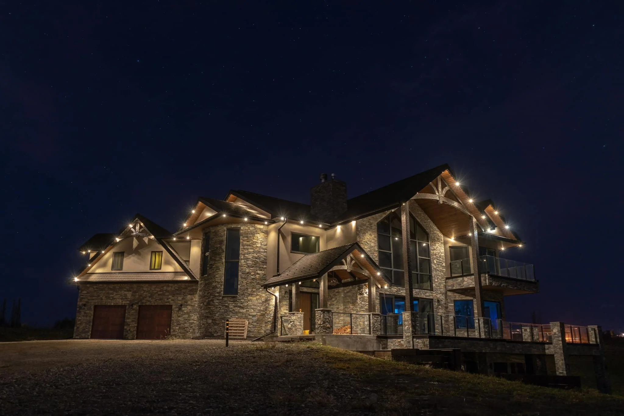 Nighttime scene of a large, upscale house made of stone and wood, with exterior lights illuminating its architecture, including balconies and a chimney, against a starry sky.