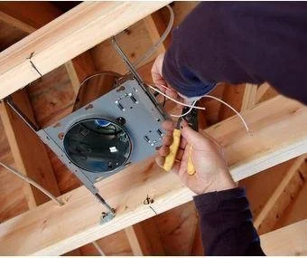 Person working on or repairing a ceiling fan or light fixture in an unfinished ceiling with wooden framing.
