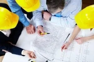 A group of five people wearing yellow hard hats gathered around a table looking at blueprints and documents.