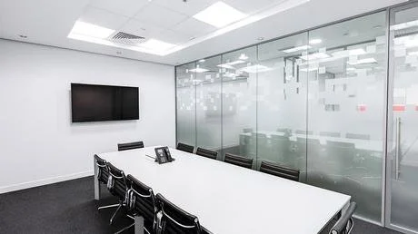 Empty conference room with a long white table, black chairs, a wall-mounted TV, and a glass partition.