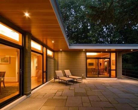 Modern house balcony with two lounge chairs, glass sliding doors, and external lighting at dusk.