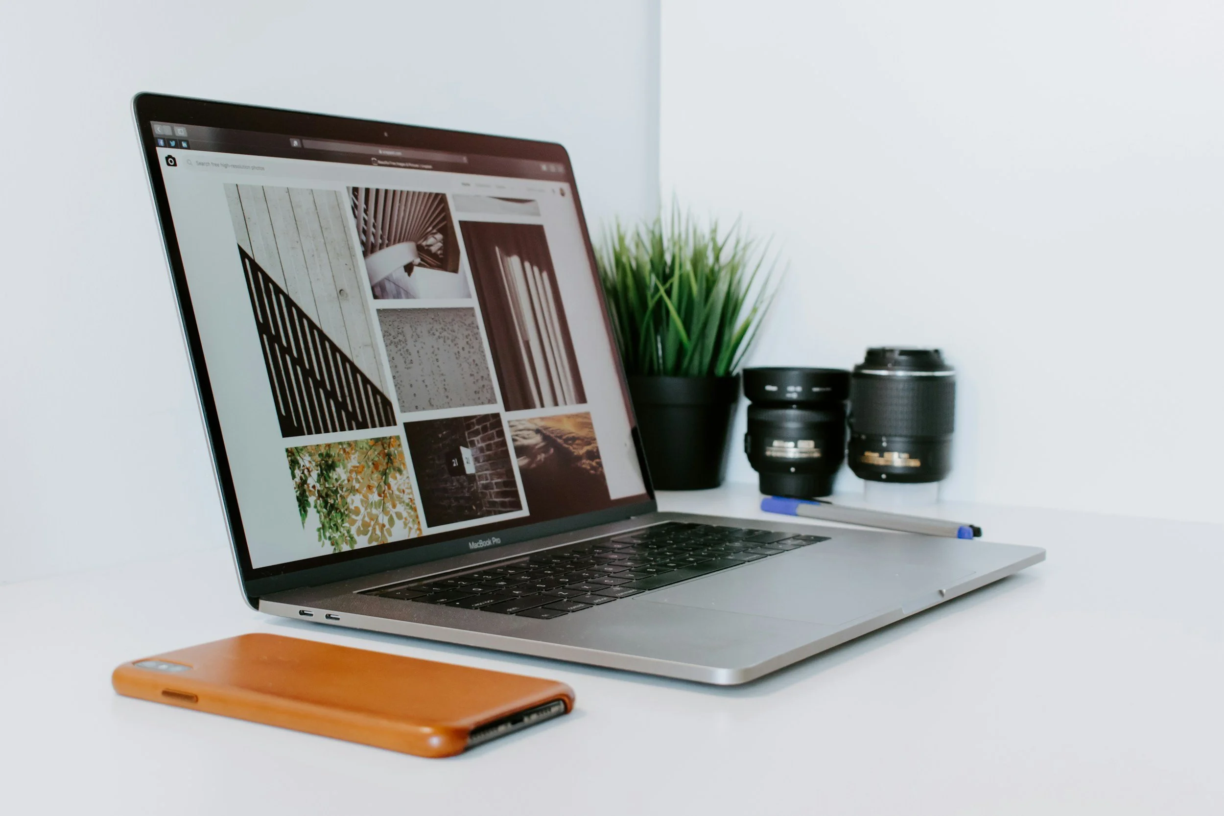 A MacBook Pro laptop on a white desk displays a grid of interior design images. To the left of the laptop is a brown phone case, and behind the laptop are a small potted plant with green grass-like leaves and two camera lenses, with a blue pen lying on the desk.