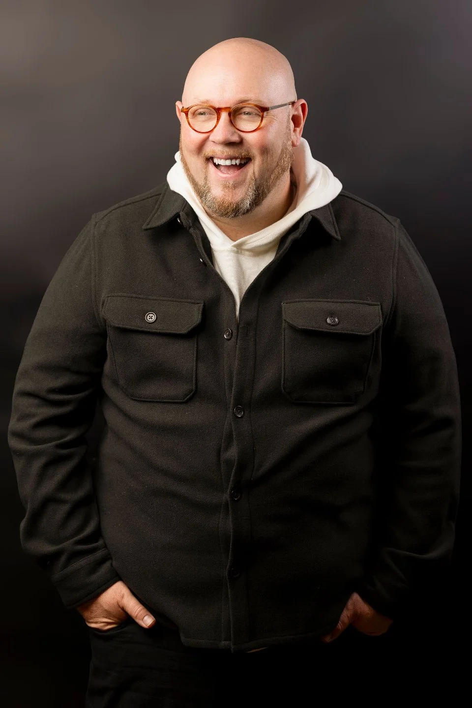A smiling man with a beard and glasses wearing a black jacket and white hoodie, standing against a dark background.