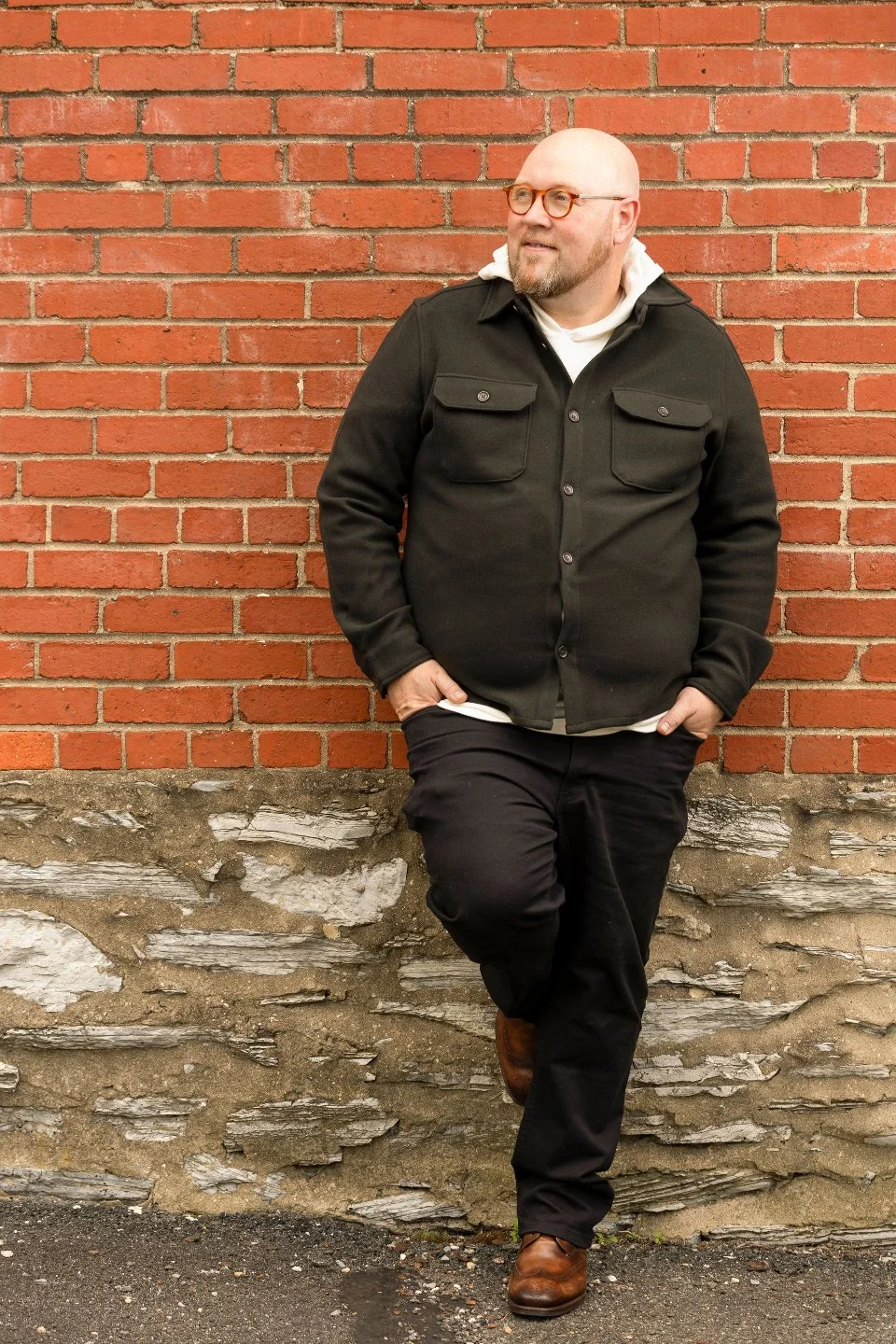 A bald man with a beard wearing glasses, a black jacket, white hoodie, black pants, and brown shoes, leaning against a brick wall outdoors.