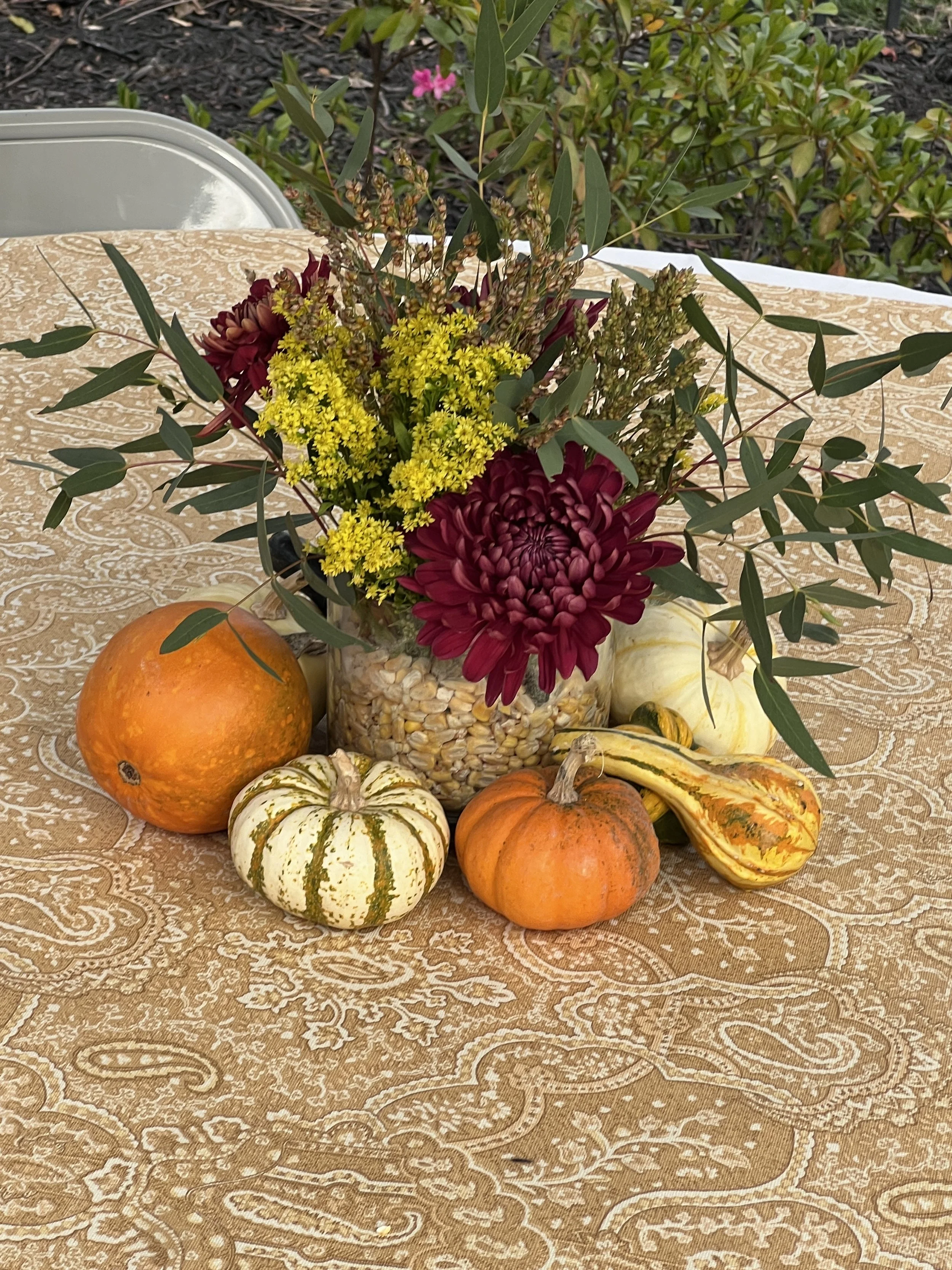 A fall-themed table arrangement with a bouquet of flowers and autumn gourds and pumpkins on a beige patterned tablecloth.